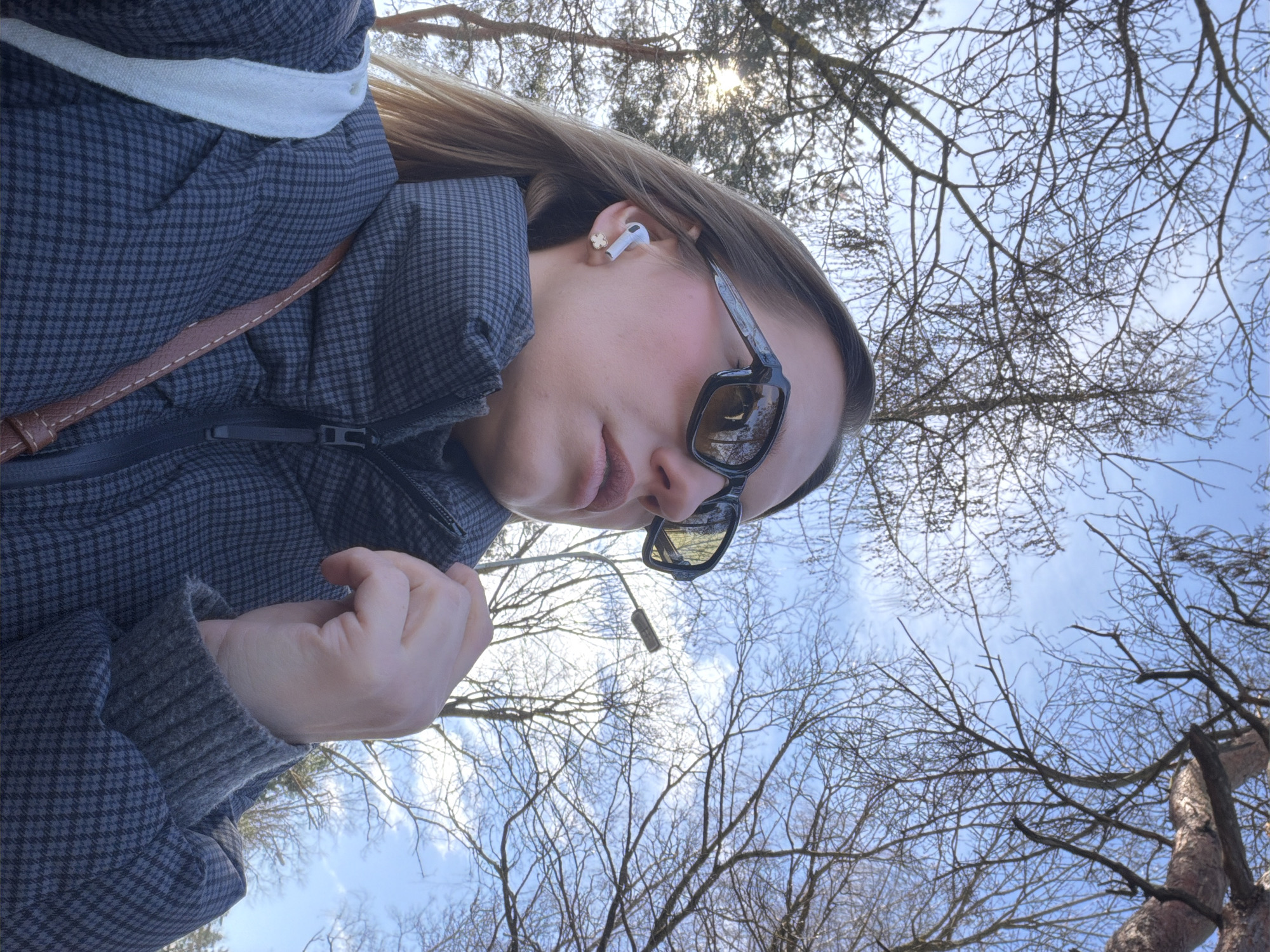 A woman wearing a gray checkered puffer jacket and sunglasses is depicted in a low-angle shot. She is wearing an earbud in her left ear, and a brown bag strap is visible on her shoulder. Her hand is near her chest. The background consists of bare tree branches against a blue sky. The photo appears to have been taken in Dreiliņi, Latvia, considering the presence of Baltic landscape features.
