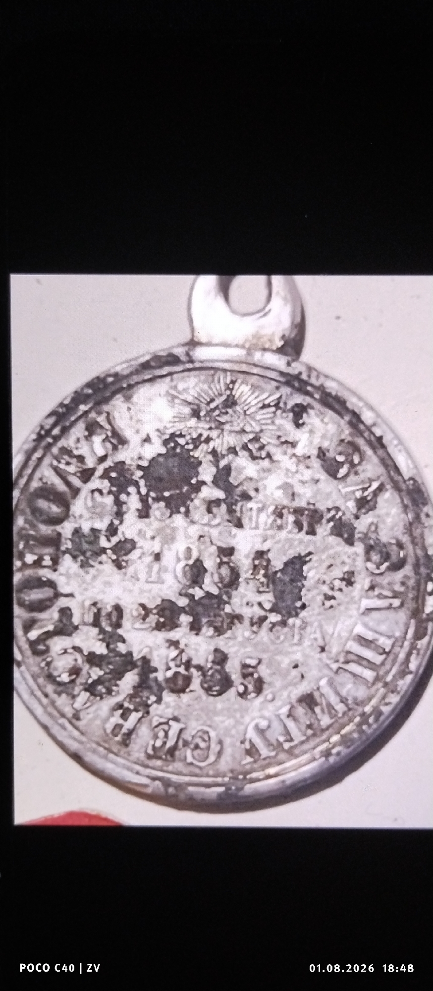 A silver-colored round pendant or medal is shown against a light background. The pendant has a raised loop at the top for attachment. The surface of the pendant is heavily textured and appears worn or tarnished, obscuring much of the detail. However, some raised lettering and imagery are discernible. A central sunburst or star-like motif is present, surrounded by circular bands of inscription. The inscription includes visible numerals and letters. In the lower portion of the image, some of the text appears to form Roman numerals, specifically "VII". There is also a red element visible at the bottom left, partially obscured by the pendant. The image also contains metadata at the bottom indicating "POCO C40 | ZV" and "01.08.2026 18:48". There is no indication or identifiable feature within the image that places it in the city of Лубянки, Russia.