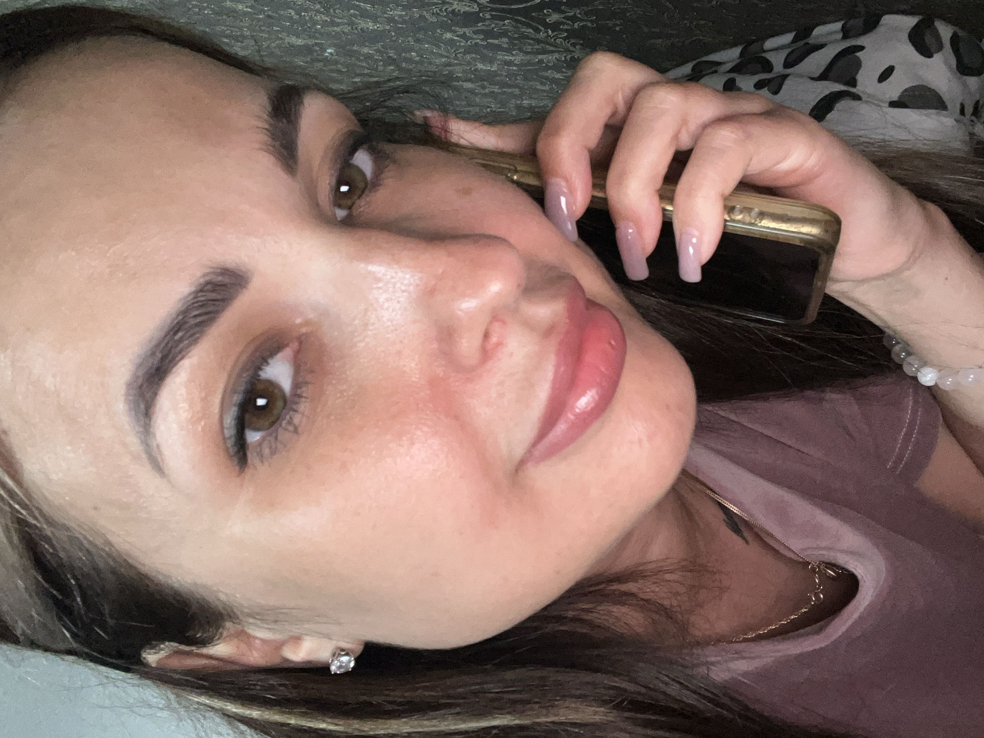 A woman is shown in a close-up, portrait-style selfie, holding a gold-colored smartphone to her right ear. She has brown hair with blonde highlights and brown eyes. She is wearing a pink shirt, a gold necklace, and a diamond stud earring in her left ear. Her fingernails are long and painted a light pink color. The background is a dark, textured wallpaper. The lighting is dim, with the primary light source appearing to be from the front. The woman's face is tilted slightly, and she appears to be speaking on the phone.