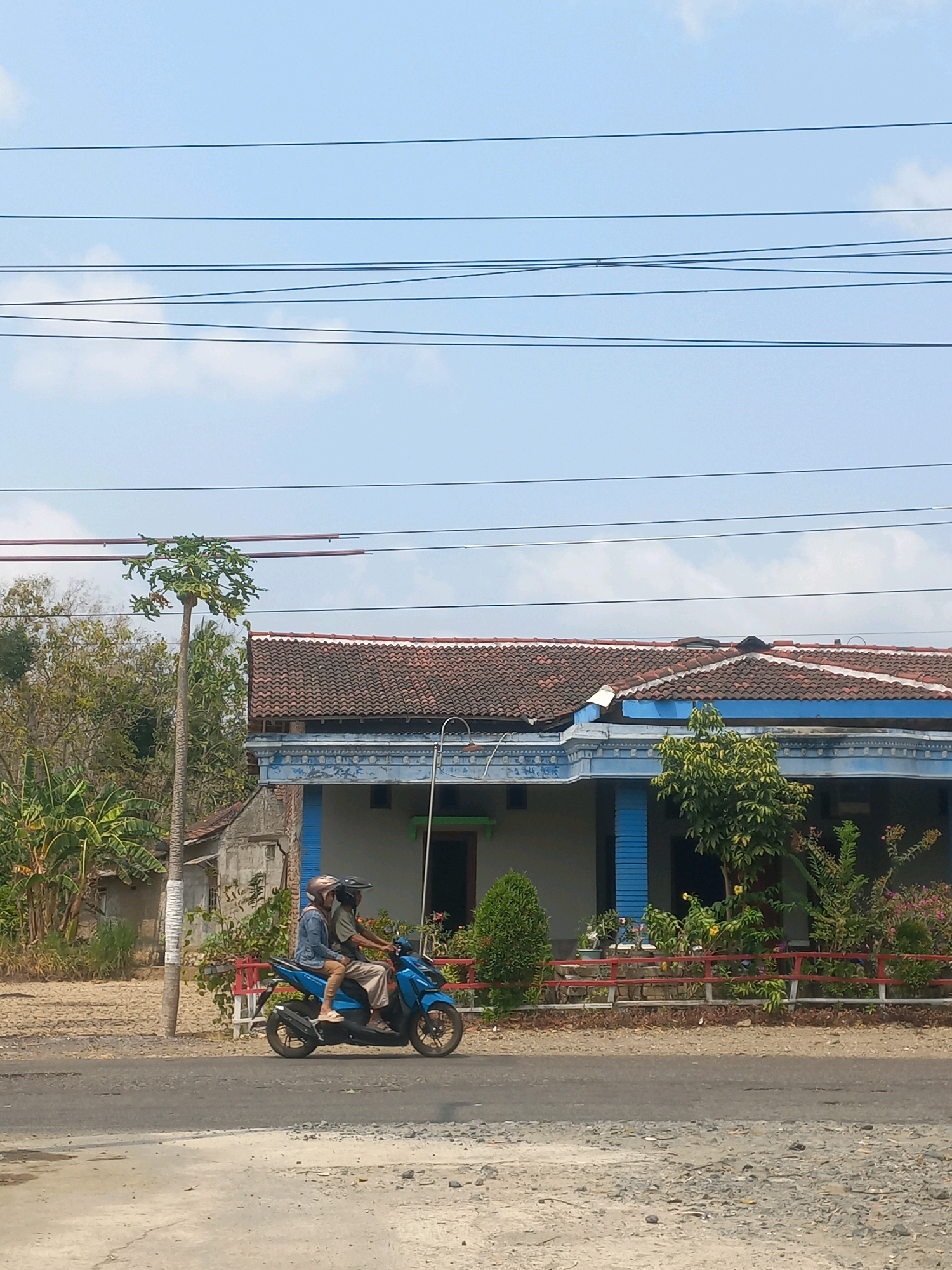 In Baturetno, Indonesia, a blue scooter carrying two individuals travels along a paved road. The rider in front, a male, wears a dark helmet, while the passenger behind him, a female, has a light-colored helmet and is dressed in a denim jacket and light pants. The scooter is passing a residential property featuring a house with a tiled roof and blue trim, surrounded by vegetation and a red-railed fence. The scene is set against a clear blue sky with thin clouds and overhead power lines. The surrounding area shows dry, cultivated land and trees.