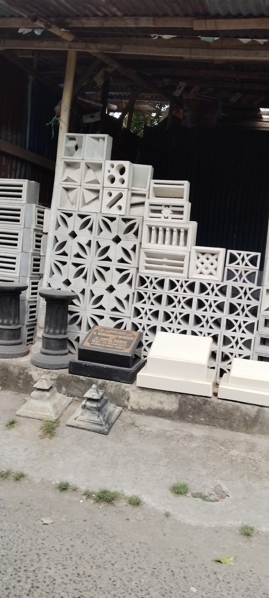 A collection of carved concrete blocks, likely decorative screens or ventilation bricks, are stacked and arranged outdoors under a weathered, corrugated metal roof supported by wooden beams. The blocks display various geometric and floral patterns. In the foreground, there are several stone pedestals of different sizes and designs, along with two dark, rectangular grave markers. One of these grave markers has an inscription in gold lettering. The ground is paved with concrete, with patches of sparse grass growing in cracks. The scene appears to be in a storage area or a workshop. The image may have been taken in the city of Mataram, Indonesia, given the context of Islamic grave markers. There are no people visible in the image.