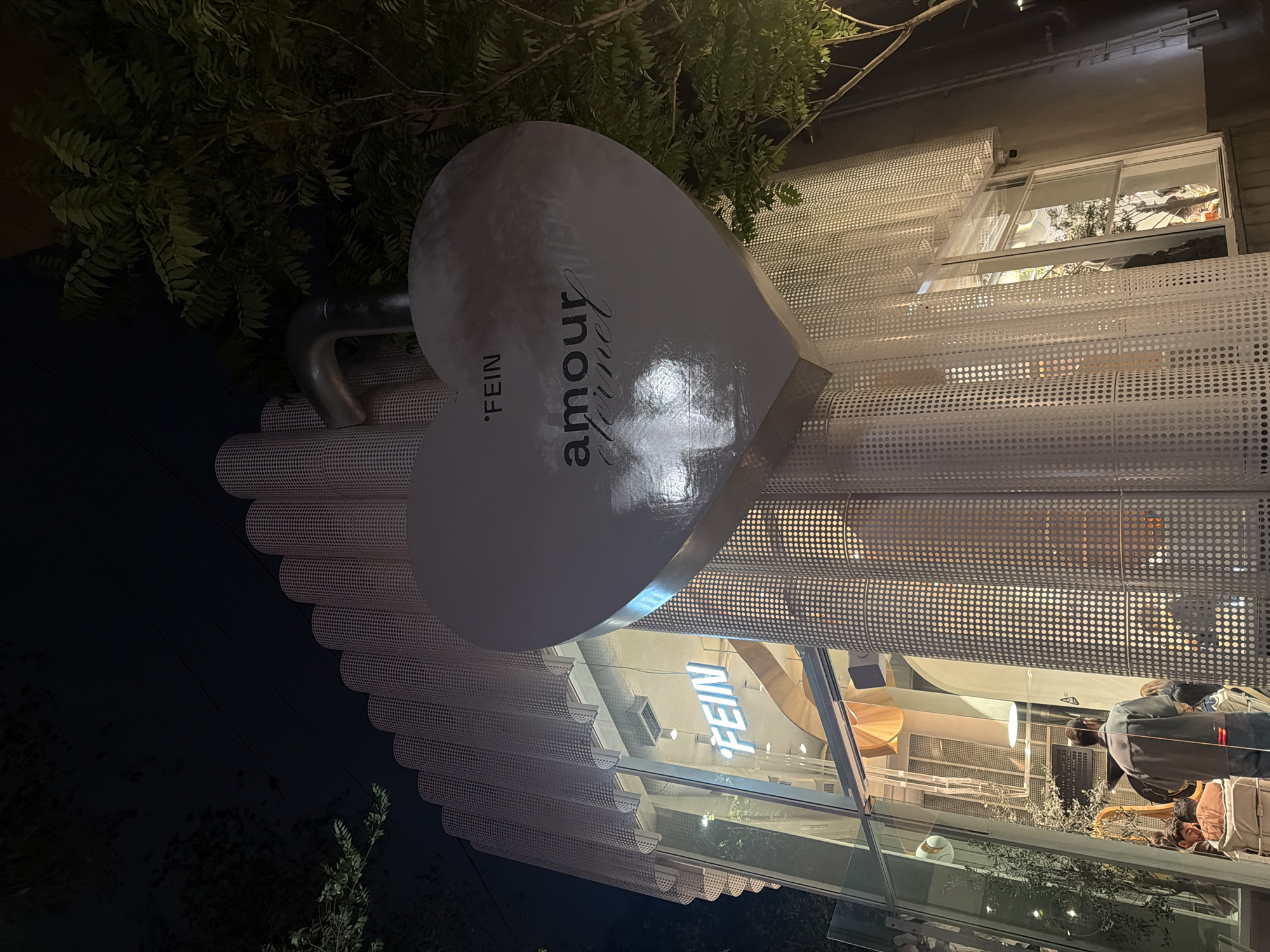 Captured in Yerevan, Armenia, a heart-shaped sign adorns the exterior of a retail store named "FEIN Amour Eternel." The sign, finished with a reflective surface, is perched above a building with a distinctive perforated facade. The facade, comprised of rows of small circular cutouts, wraps around the store, adding a layer of texture and visual interest. Inside the store, people can be observed, their reflections visible in the glass windows. The environment surrounding the store is lush, with greenery and trees in the foreground. The lighting indicates it is nighttime, and the sky is dark.