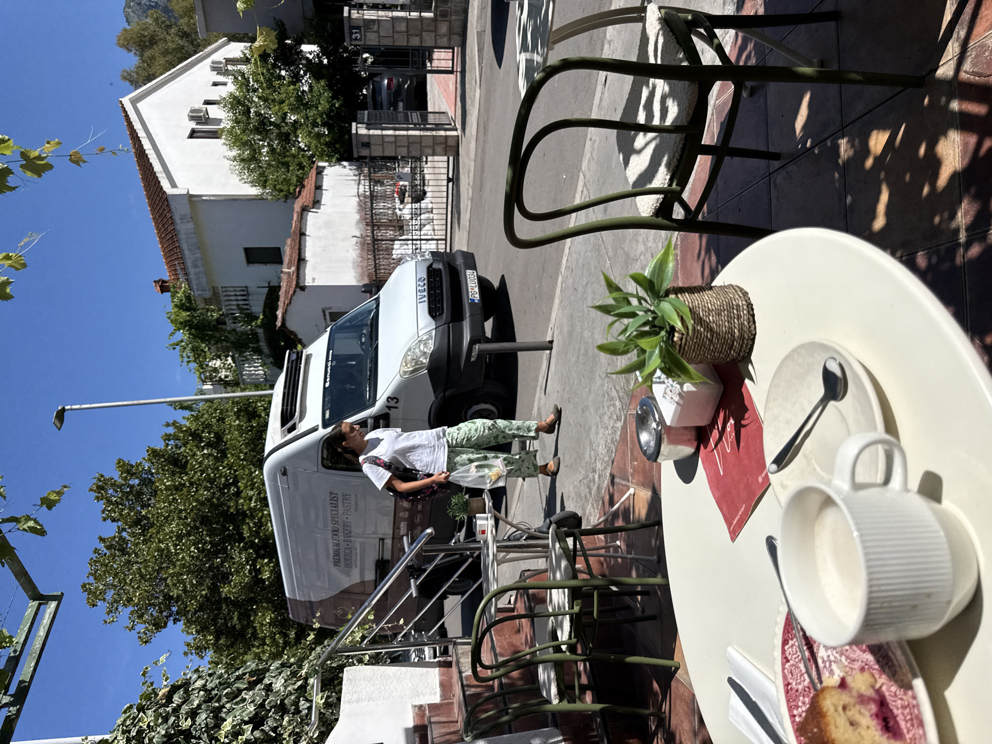 A woman walks past a cafe in Budva, Montenegro. She is carrying a white plastic bag in her right hand and has a backpack on her left shoulder. The woman is wearing a white t-shirt and patterned green pants. She is walking on the sidewalk in front of a cafe with outdoor seating. There are several tables and chairs in the foreground, and a white van is parked on the street behind the woman. The van has the words "PREMIUM FOOD SPECIALIST HORECA BAKERY PASTRY" written on its side. In the background, there is a white building with a red tile roof and trees. The sky is clear and blue.