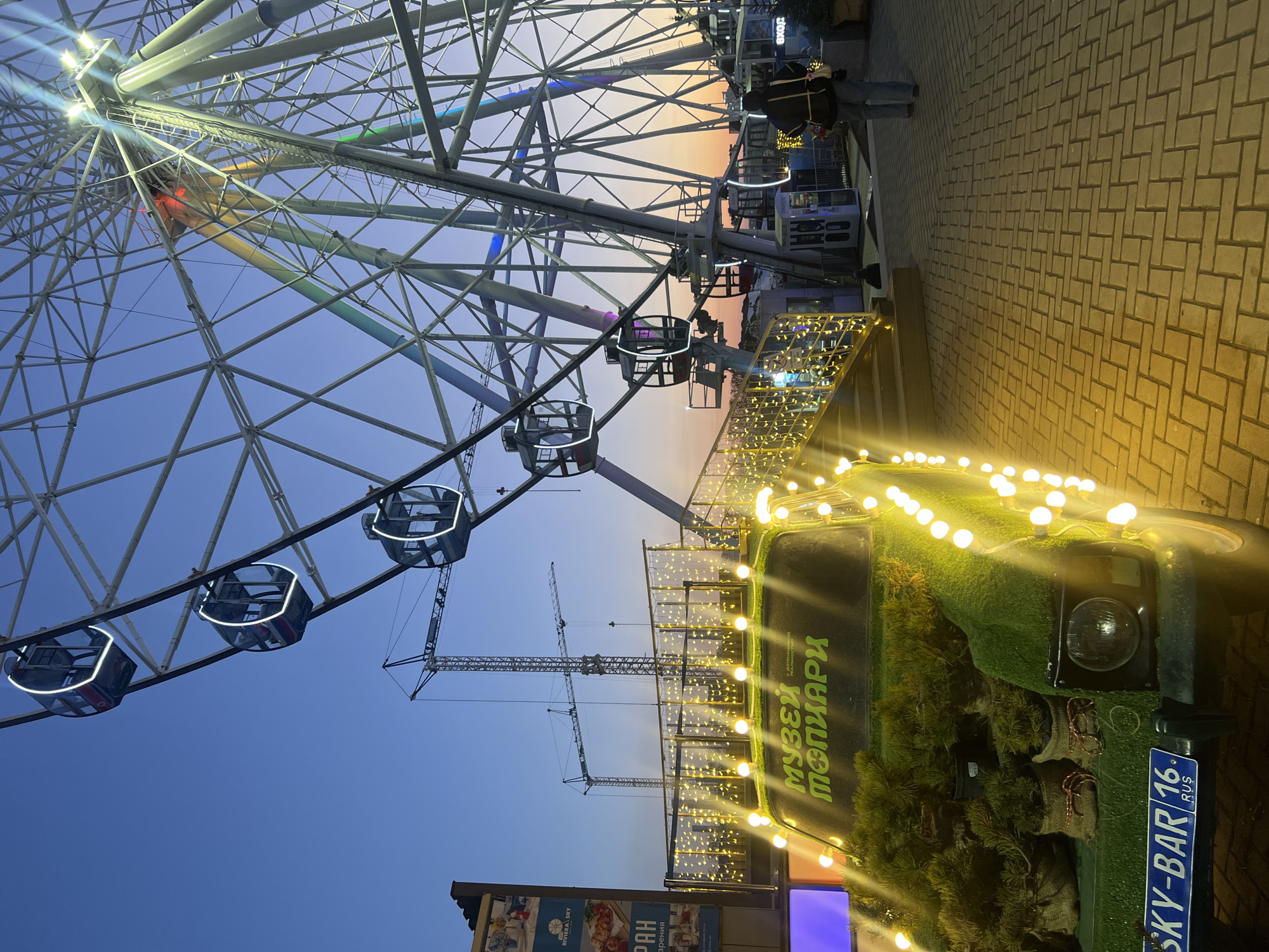 The image shows a Ferris wheel against a blue and orange twilight sky. The Ferris wheel's structure is made of white metal with enclosed, lit-up gondolas. In the background, cranes indicate nearby construction. In the foreground, an unusual vehicle decorated with fake grass and string lights is visible. The vehicle's license plate reads "SKY-BAR 16 RUS," and the side has text in Cyrillic characters. The scene is located in Kazan, Russia.
