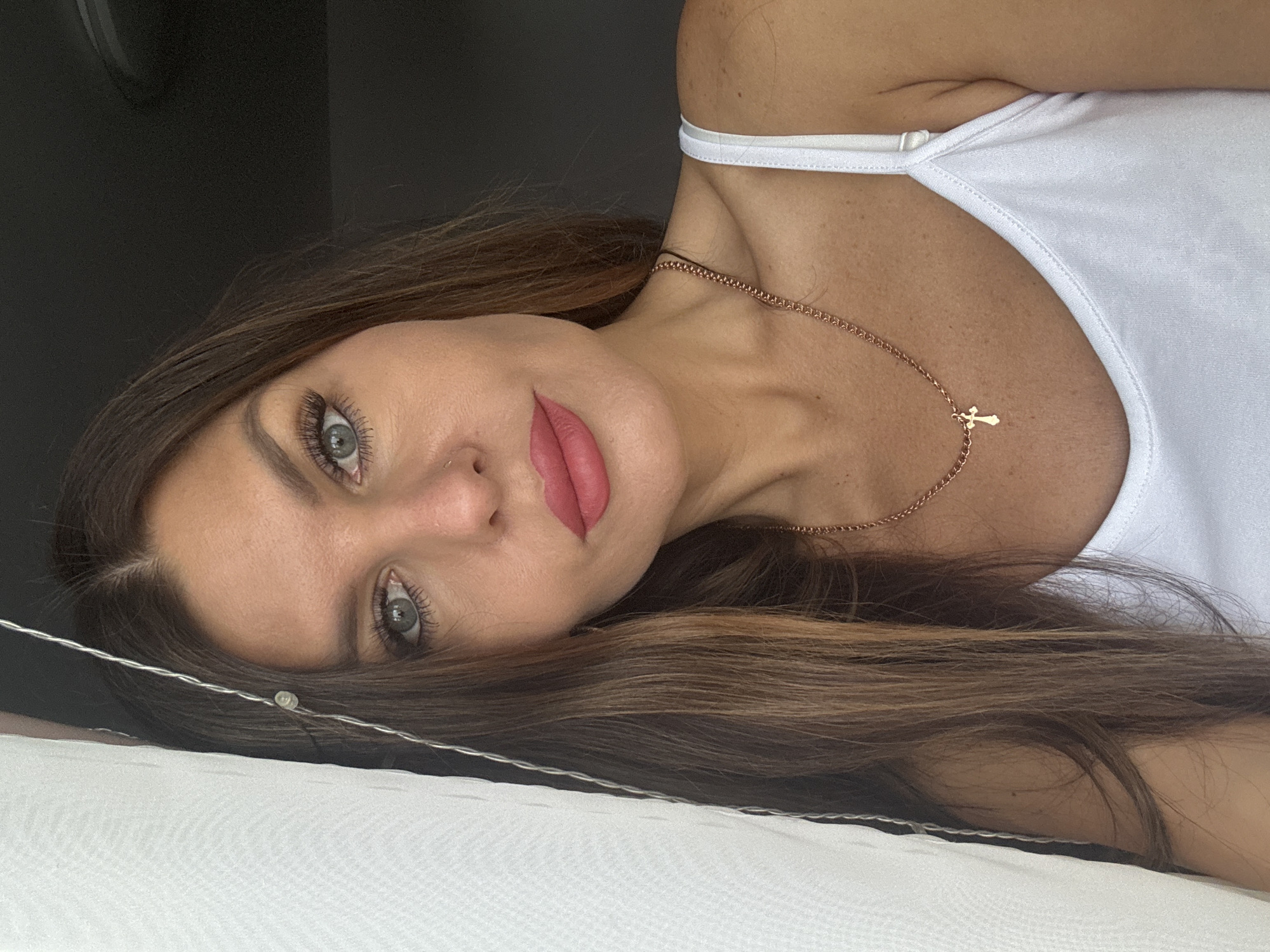 A woman with long brown hair and blue-gray eyes is lying down, presumably on a bed with white sheets. She is wearing a white tank top and a gold necklace with a small cross pendant. Her makeup includes noticeable mascara and pink lipstick. Part of a string of fairy lights is visible near her face. The location might be inferred from the prompt as the city of Chaikovsky, Russia, but there are no elements in the image itself that confirm this. The background is a dark solid color, possibly a wall.