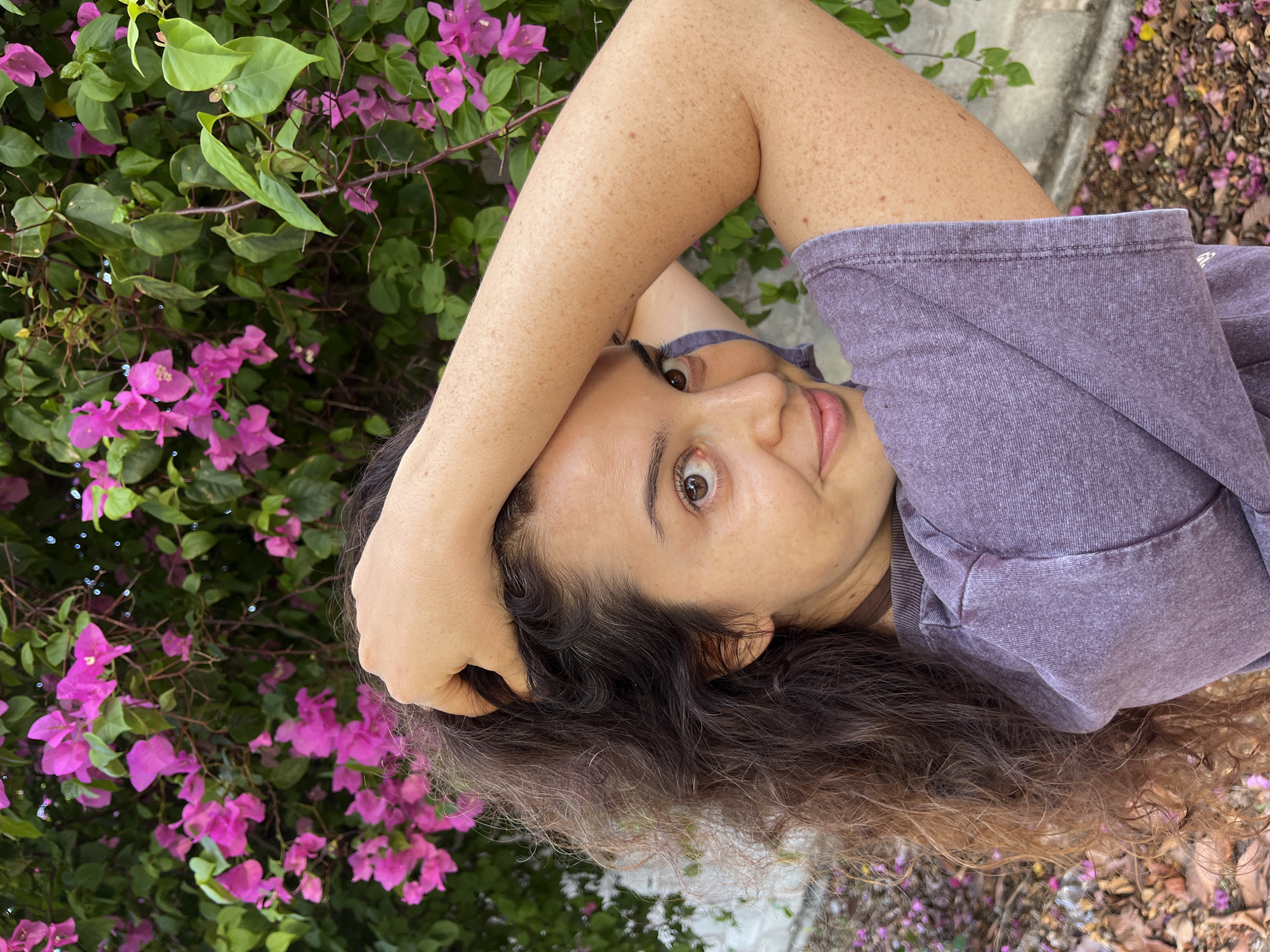 A young woman with long, curly brown hair and brown eyes smiles as she poses with her arm behind her head in front of a bush with pink bougainvillea flowers. The woman is wearing a purple t-shirt. The background shows the flowers and green leaves of the bush, along with a blurry, light-colored wall and the ground covered in fallen leaves and small pink flowers. The scene appears to be outdoors, possibly in a garden or park.