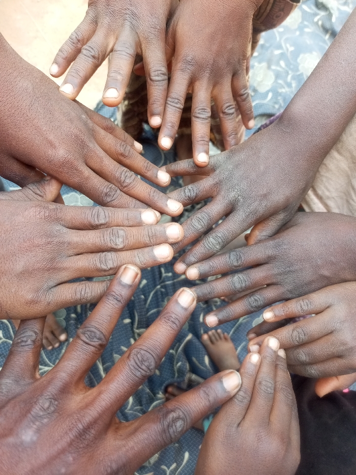 An overhead view captures a central cluster of approximately ten dark-skinned hands, palms down, with fingers splayed and overlapping. The hands exhibit varying sizes, suggesting a mix of adult and juvenile participants. Fingernails are visible, uniformly unpolished, light beige to off-white, with some displaying minor subungual debris. Beneath the hands, a dark fabric featuring a repeating light floral or leaf pattern is predominantly visible, alongside sections of plain light-toned fabric. In the lower frame, two bare feet of an infant or young child rest upon the patterned fabric, with a third smaller foot partially visible. The upper background reveals a light, possibly sandy or concrete, ground surface. The scene is located in Tempure, Nigeria.