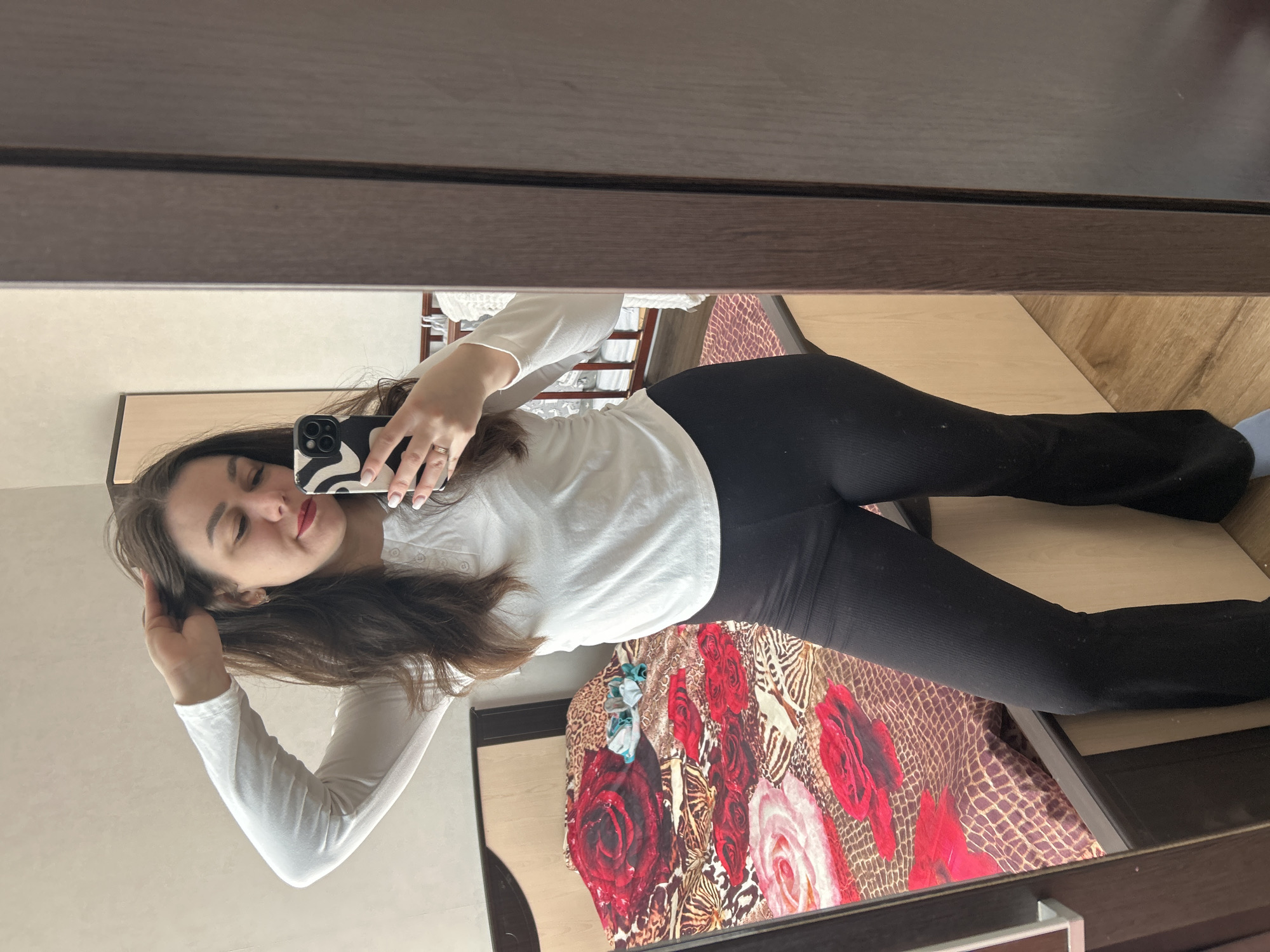 The image appears to be a selfie taken in a room. A woman with long brown hair is holding a phone with a black-and-white patterned case in her right hand, presumably to take the picture. She is wearing a white long-sleeved shirt and black leggings. The room appears to be a bedroom, with a bed visible in the background featuring a colorful floral and animal print comforter. The floor is wooden. The photo is taken in a mirror, with part of the mirror's wooden frame visible at the top and bottom. The metadata doesn't include the location. The assertion of the location as Chaikovsky, Russia is unfounded without supporting evidence.