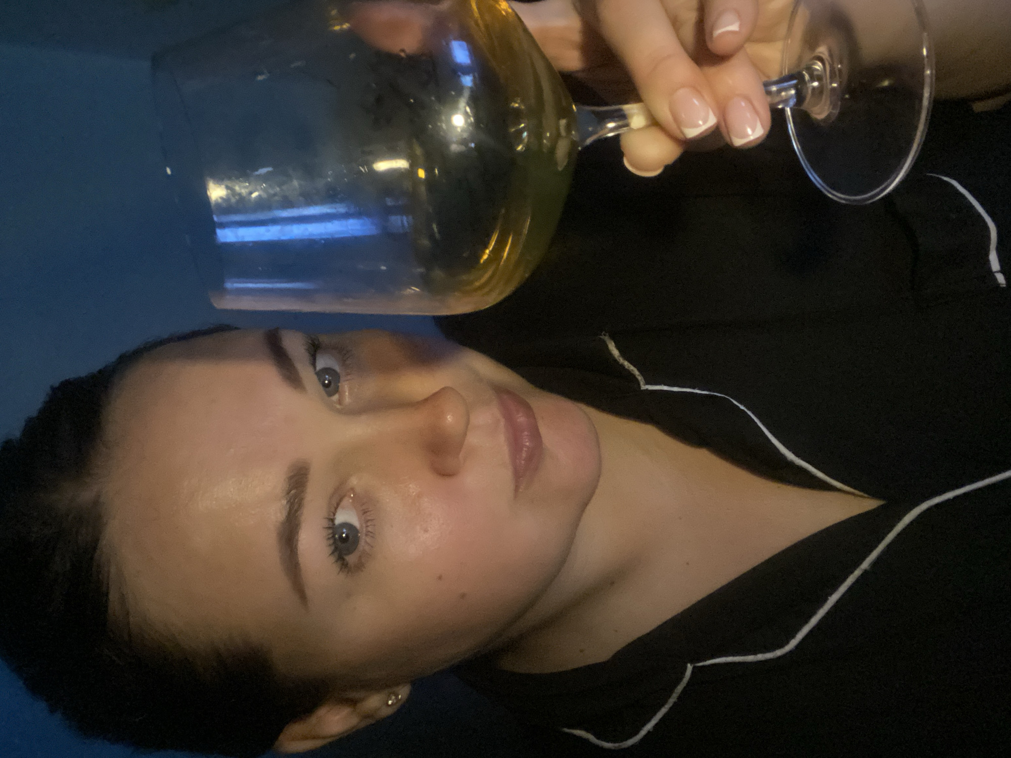 A woman is pictured holding a glass of pale liquid (likely wine) in front of her face. She has fair skin, light-colored eyes, and dark hair pulled back. She wears a dark shirt with white piping. The image appears to be taken indoors. There is no information to indicate the location is Sochi, Russia.
