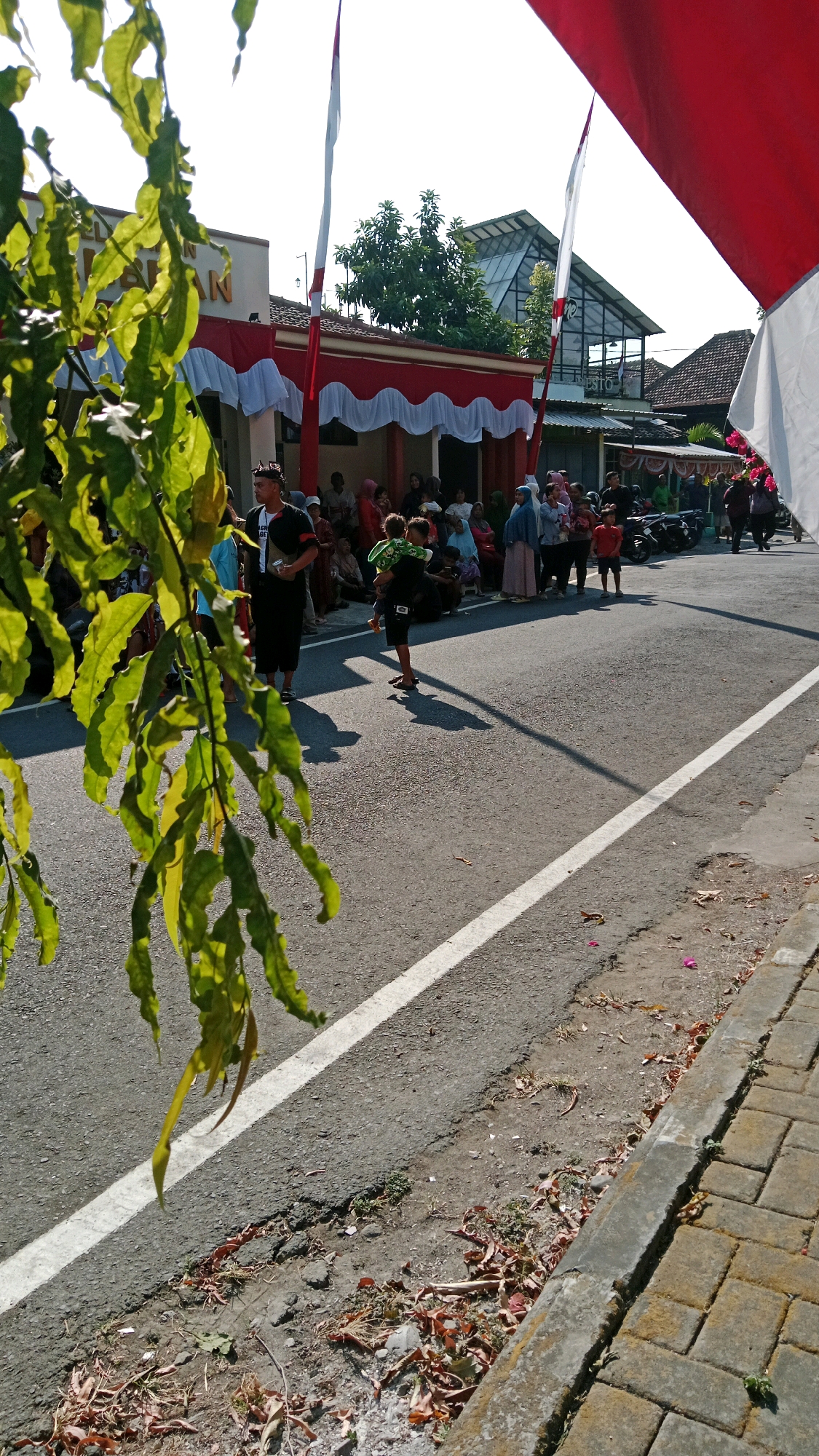 The image captures a street scene in Magetan, Indonesia, likely during a public gathering or celebration, as indicated by the presence of Indonesian flags (red and white) and a crowd of people lining the street. The foreground features a large branch with green leaves, partially obscuring the view. The street itself is paved, with a white line marking the lane. On the right side of the street, a brick sidewalk is visible.
In the background, a building with red and white decorative trim is prominent, suggesting it might be a public institution or a venue for an event. People of various ages are gathered, some standing and others sitting, appearing to watch something or await an event. Several individuals are dressed in traditional clothing. One man in the middle ground is holding a child, while other adults are engaged in conversations or observing their surroundings. Motorbikes are parked along the side of the road. The overall atmosphere appears to be sunny and outdoor.