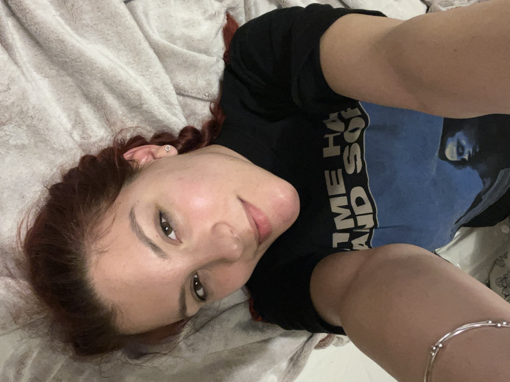 A woman with auburn hair braided on one side and wearing a black t-shirt with faded blue and white lettering is lying on her back on a light gray blanket. She is looking at the camera. The scene appears to be indoors. There is no visible information to determine the location in the city of Sofia, Bulgaria.