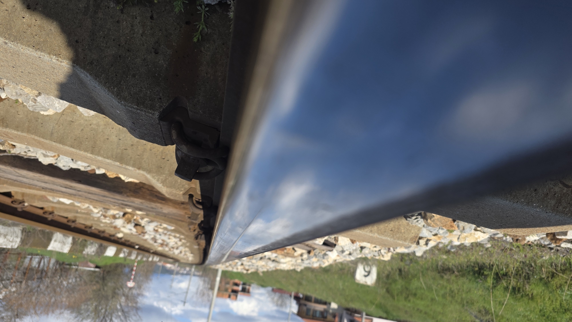 The image depicts a close-up view of a railway track near Blagoevgrad, Bulgaria. The focus is on the rail itself, showing the metal surface reflecting the sky above. The rail is secured with fastening clips onto a concrete tie. Ballast, consisting of crushed rock, fills the space between the concrete ties. In the background, vegetation and buildings are visible. A sign with the number '9' is on the ground.