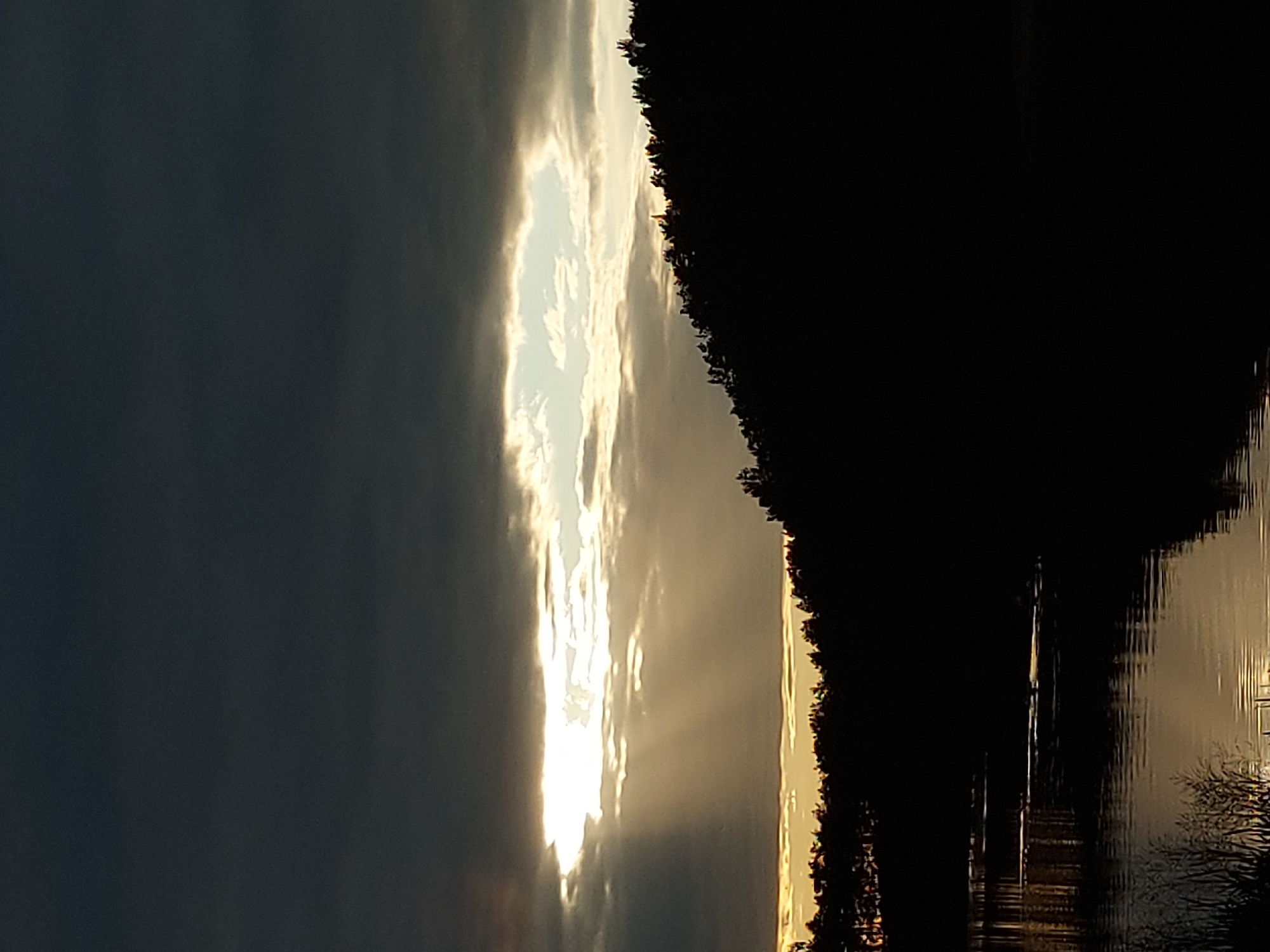 A dark, cloudy sky looms over a silhouetted forest that slopes down to a body of water. Sunlight streams through a break in the clouds, casting visible rays onto the scene. The water reflects the light and the dark forest. The image depicts an outdoor, natural environment.