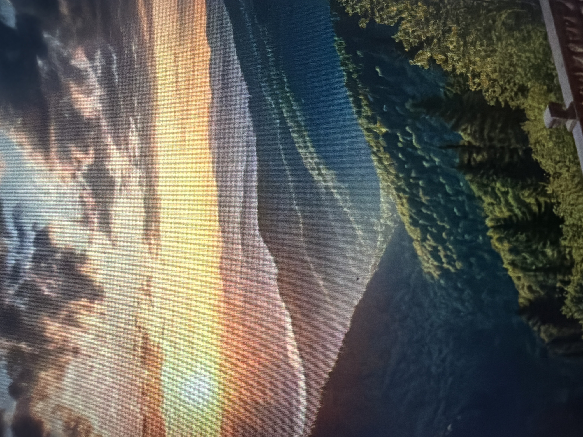 The image is a landscape photograph of rolling mountains at sunrise or sunset. The sky is filled with dramatic clouds in shades of orange, yellow, and dark gray. The sun, a bright orb, is visible on the left side of the frame, casting a warm glow across the sky. Layers of mountains recede into the distance, with each layer appearing a lighter shade of blue and purple due to atmospheric perspective. The closest mountains are covered in dense green forest. In the foreground, a wooden railing is visible, suggesting this view is from an overlook or balcony. The image quality appears to be slightly pixelated, possibly due to being a photograph of a screen.
The image does not depict the city of Baan Mae Nam, Thailand. The landscape is characteristic of mountainous regions, not the geographical features typically found in Baan Mae Nam, which is located in a valley.