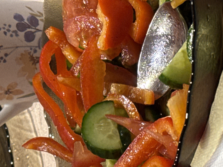 A close-up shot captures a vibrant salad, featuring sliced red bell peppers and green cucumbers. A silver spoon is visible, partially obscuring the view. The salad is contained within a black bowl adorned with small white dots, resting on a surface with a floral pattern. The scene is illuminated, casting highlights on the ingredients, emphasizing the fresh, colorful presentation.