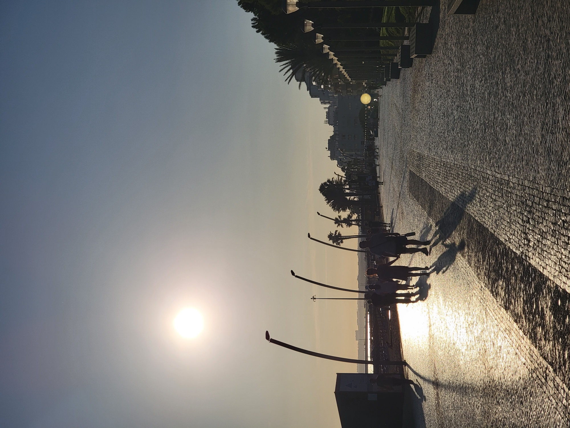 This is a photograph of the promenade in Quarteira, Portugal, at sunset. The sun is low in the sky, casting a warm glow and long shadows across the cobblestone pavement. Several people are walking along the promenade, silhouetted against the bright sky. Lampposts with distinctive curved arms line the promenade, interspersed with palm trees and benches. In the background, buildings and a distant shoreline are visible. The sea is to the left of the frame, with the sunlight reflecting off its surface.