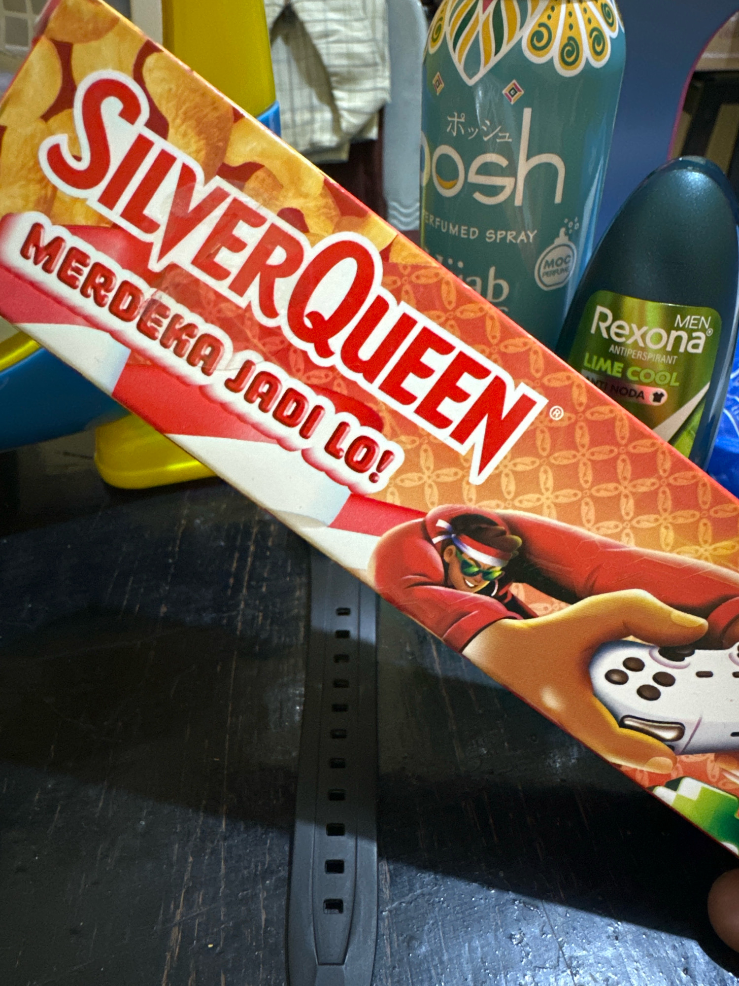 A close-up, overhead shot captures a table with several items, including a Silver Queen chocolate bar, a Posh perfumed spray, and a Rexona men's antiperspirant. The Silver Queen bar is prominently displayed, with its red and white packaging featuring the brand name in large white letters. Below "Silver Queen" are the words "MERDEKA JADI LO!" The Posh spray can is teal with decorative patterns and text in Japanese and English, including "POSH" and "PERFUMED SPRAY." The Rexona antiperspirant is dark green with a lime green label indicating "MEN ANTIPERSPIRANT LIME COOL." A black watch strap is visible across the bottom of the frame, and a person's finger is touching the right edge of the image, suggesting they are holding or interacting with the items. The background appears to be a wooden surface with a textured, slightly worn appearance. The setting does not provide any specific clues to the city of Saruni, Indonesia.