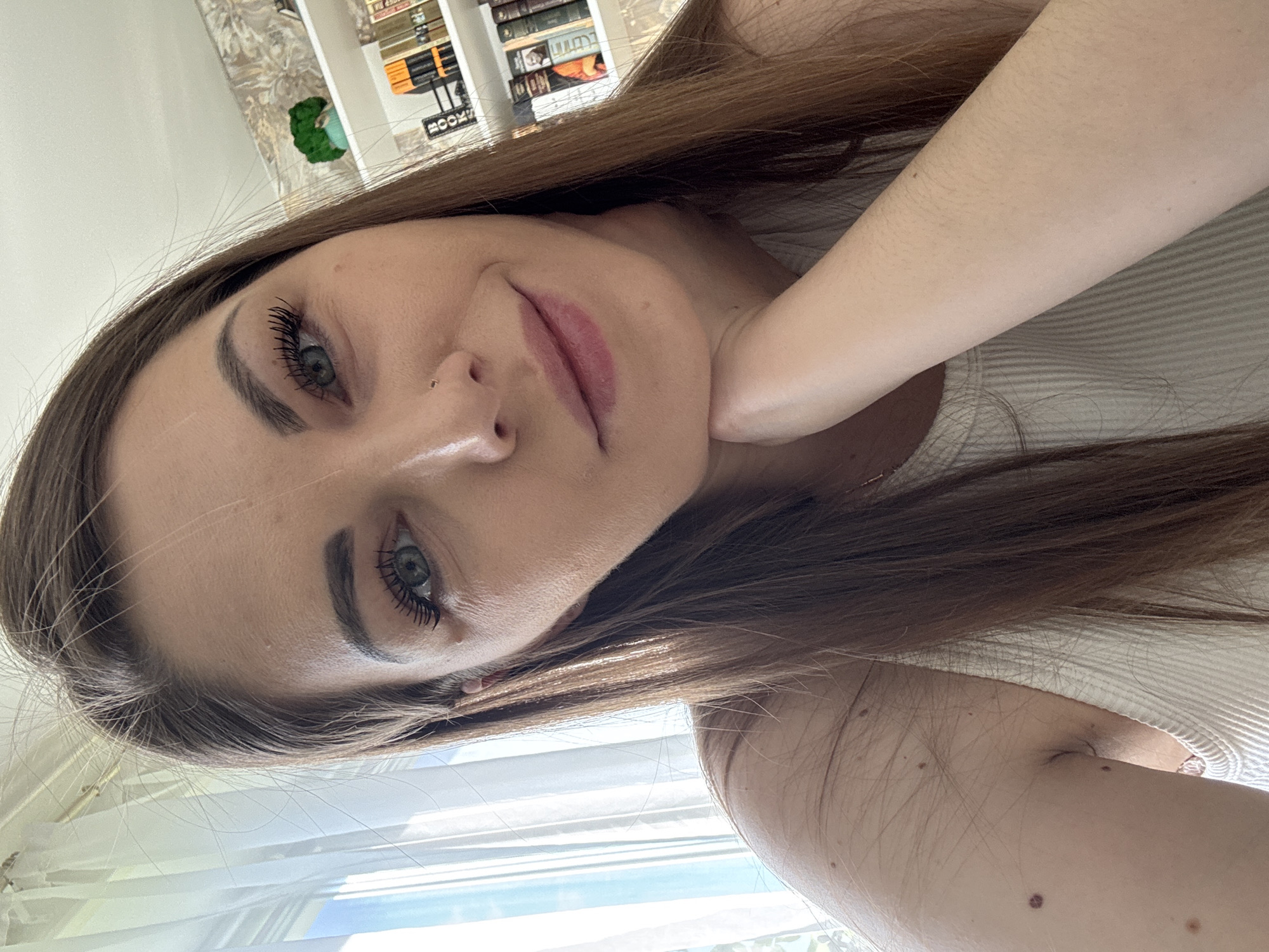 A close-up, slightly angled, portrait shot of a woman with long, brown hair. She has fair skin, dark eyebrows and eyelashes, and light-colored eyes. She is wearing a white top. In the background, blurred, is a room with bookshelves and a window with blinds. The location information (Раздолье, Russia) is not clearly visible or suggested by the image's content.