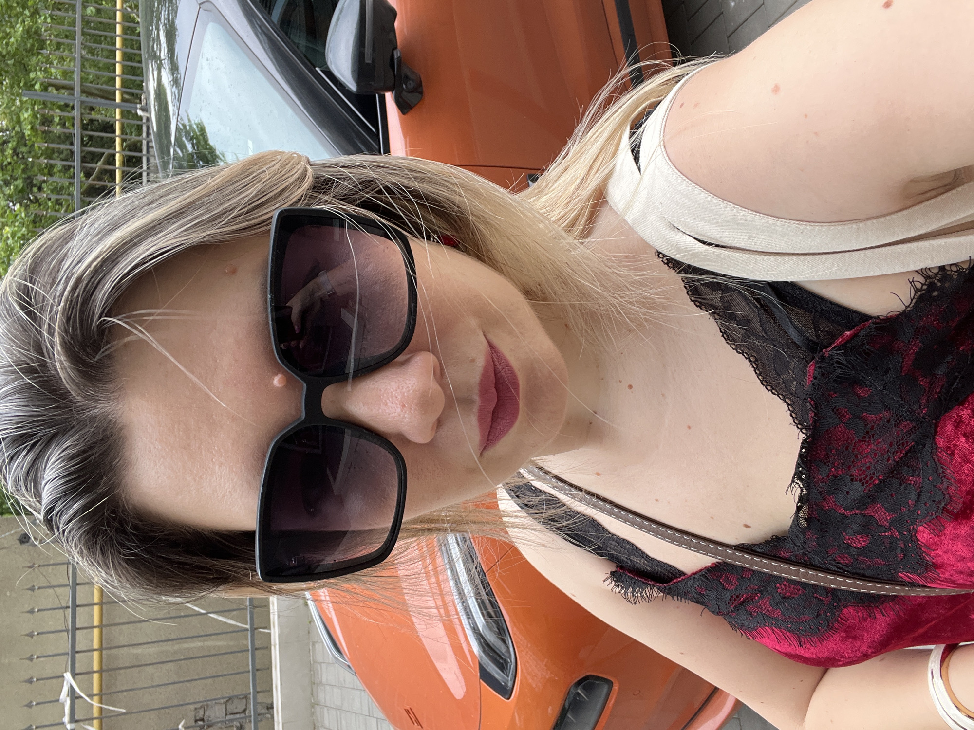 A woman is taking a selfie in front of an orange car parked outdoors. She is wearing large black sunglasses, a red and black lace top, and a light-colored strap from a bag. Her blonde hair is partially obscuring her face. The background shows a metal fence and greenery, suggesting an urban or suburban setting. There is no specific indication that the location is Sochi, Russia.