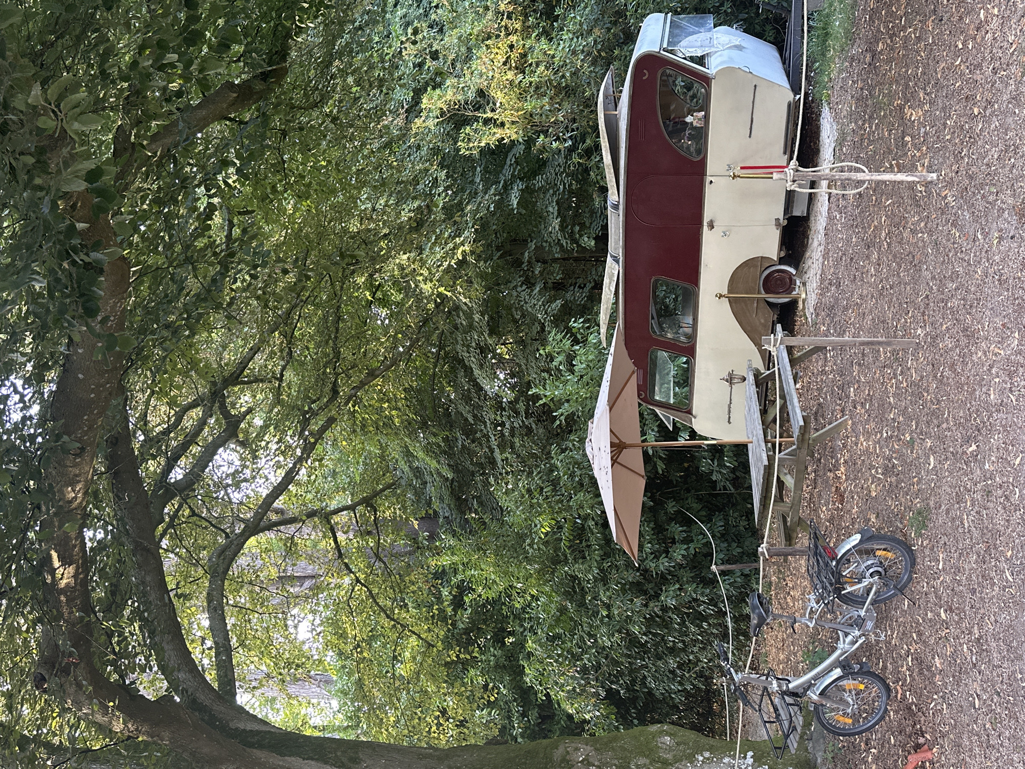 This image captures a tranquil outdoor scene, likely a campsite, nestled within a lush green environment characteristic of Saint-Vaast-du-Val, France. A vintage caravan, predominantly cream-colored with a deep red roof and accents, is parked on a bed of fallen leaves and mulch. The caravan's design suggests it is from an earlier era, featuring curved windows and a polished exterior.
In the foreground, a silver folding bicycle with front and rear baskets is parked, its wheels resting on the leaf-covered ground. A picnic table made of weathered wood, accompanied by an open, light-colored umbrella, is set up near the caravan, suggesting a space for relaxation and dining. The area is delineated by simple rope barriers supported by wooden posts.
Behind the caravan and picnic table, dense foliage and trees form a natural backdrop, with sunlight filtering through the leaves, creating dappled light and shadow. The overall impression is one of a peaceful, rustic retreat, bathed in the soft light of late afternoon or early morning. No people are visible in the frame, emphasizing the solitude and quietude of the location. The environment is natural and unmanicured, with a sense of undisturbed natural beauty.