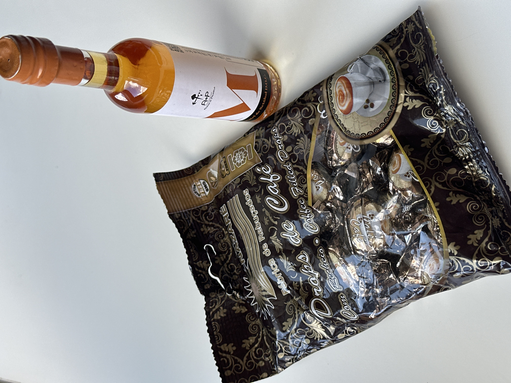 A bag of coffee-filled drops and a small bottle of amber liquid are displayed on a white surface. The bag is dark brown with ornate gold patterns and features images of coffee cups. The label on the bag reads "Drops de Café" and "Coffee Filled Drops." The bottle has a white label with a prominent orange letter "M" and a logo that appears to be "R+P ADEGA D' PALMELA." The liquid in the bottle is translucent and appears to be a fortified wine, possibly Moscatel de Setúbal, given the text on the label. The background is a plain white surface, likely a table or countertop, with soft lighting creating subtle shadows. The scene is static, with no people or actions depicted. The photograph was taken from a slightly elevated angle, focusing on the products. The location is not specified in the image or its context.