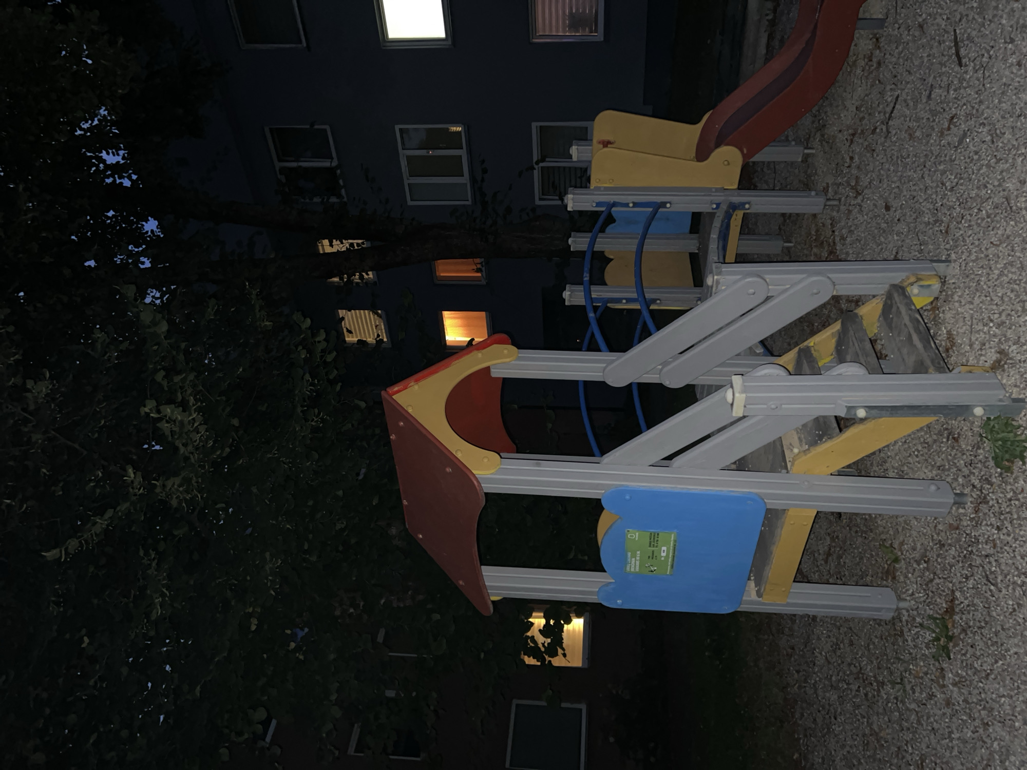 A nighttime view of a children's playground structure in Vilnius, Lithuania, is presented. The playground is situated on a gravel surface, with a dark-colored building visible in the background. The building has several windows, some of which are illuminated, suggesting occupancy. A tree with dark foliage stands prominently in front of the building, partially obscuring the view. The playground structure itself is colorful, featuring red, yellow, and blue elements, and includes a slide. The scene is dark, typical of nighttime conditions, with artificial light emanating from the building's windows and possibly some ambient light from the surrounding area. There are no people visible in the image, and no discernible actions or interactions are taking place. The overall impression is one of stillness and quiet.