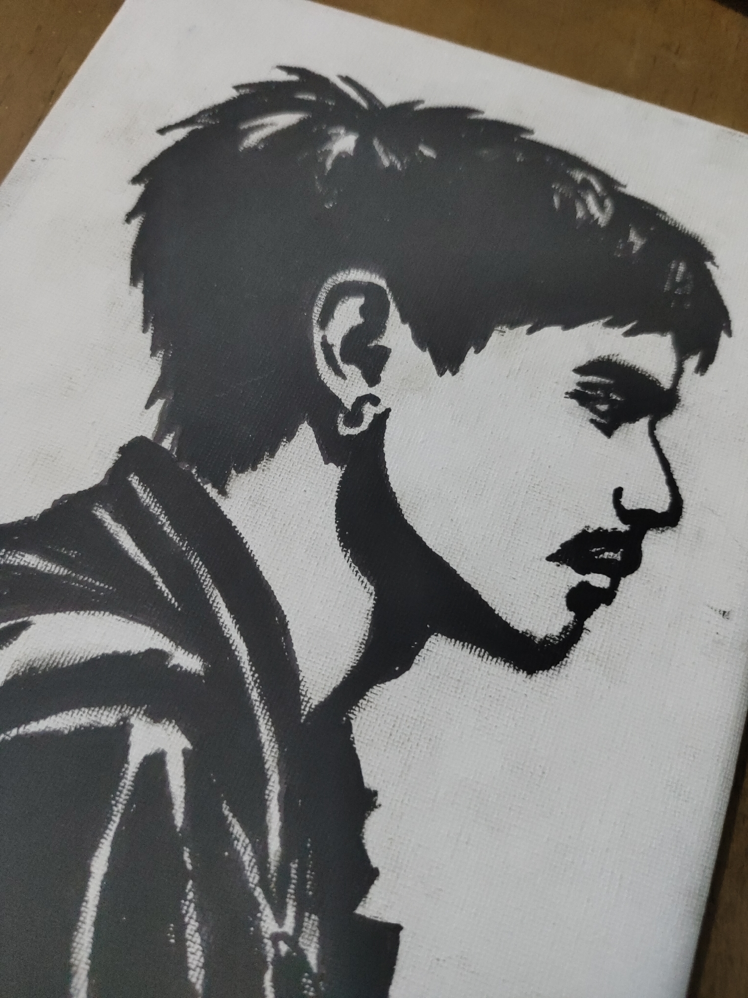 The image displays a high-contrast, black and white portrait of a male subject rendered on a coarse white canvas, indicative of a stencil, screen print, or high-contrast graphic art. The artwork is located in Davao City, Philippines.
The subject is depicted in a right-facing profile, occupying the central and left portions of the canvas. He possesses short, textured black hair styled upwards, and a prominent ear featuring a small, circular hoop earring. His eye is represented by a dark, almond shape beneath a low brow. A defined nose, a short mustache, and full, dark lips are clearly visible. His jawline is strong and angular, leading to a distinct chin. The subject wears a collared garment, possibly a jacket or shirt, with vertical lines or folds suggesting texture on the left shoulder and arm area.
The background is uniformly white, allowing the black silhouette of the subject to stand out sharply. The canvas material exhibits a noticeable woven texture throughout the white areas and partially through the black inked sections, indicating the physical medium of the artwork. The edges of the canvas are visible, suggesting it is an unframed piece.