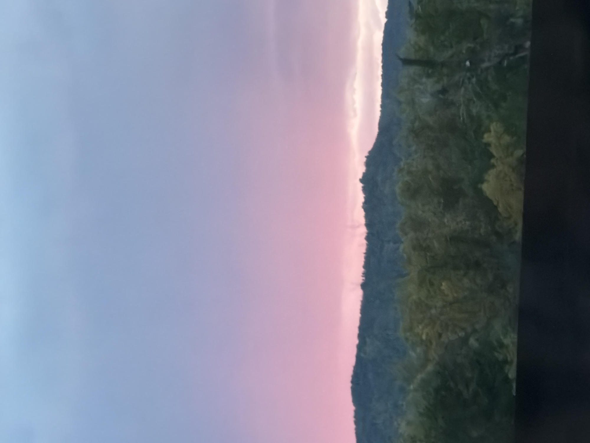 The image depicts a natural landscape, likely at dusk or dawn, given the soft pastel colors in the sky. The sky transitions from a pale blue at the top to a vibrant pink and purple gradient towards the horizon. Below the sky, a dark, tree-covered hillside forms a prominent silhouette. In the foreground, there is a dark, horizontal band, possibly a railing or a ledge, obscuring the very bottom of the scene. The overall impression is one of natural beauty and tranquility. There are no visible people, animals, or man-made structures apart from the implied viewpoint from which the photograph was taken. There is no information in the image to suggest it is located in Samara, Russia.