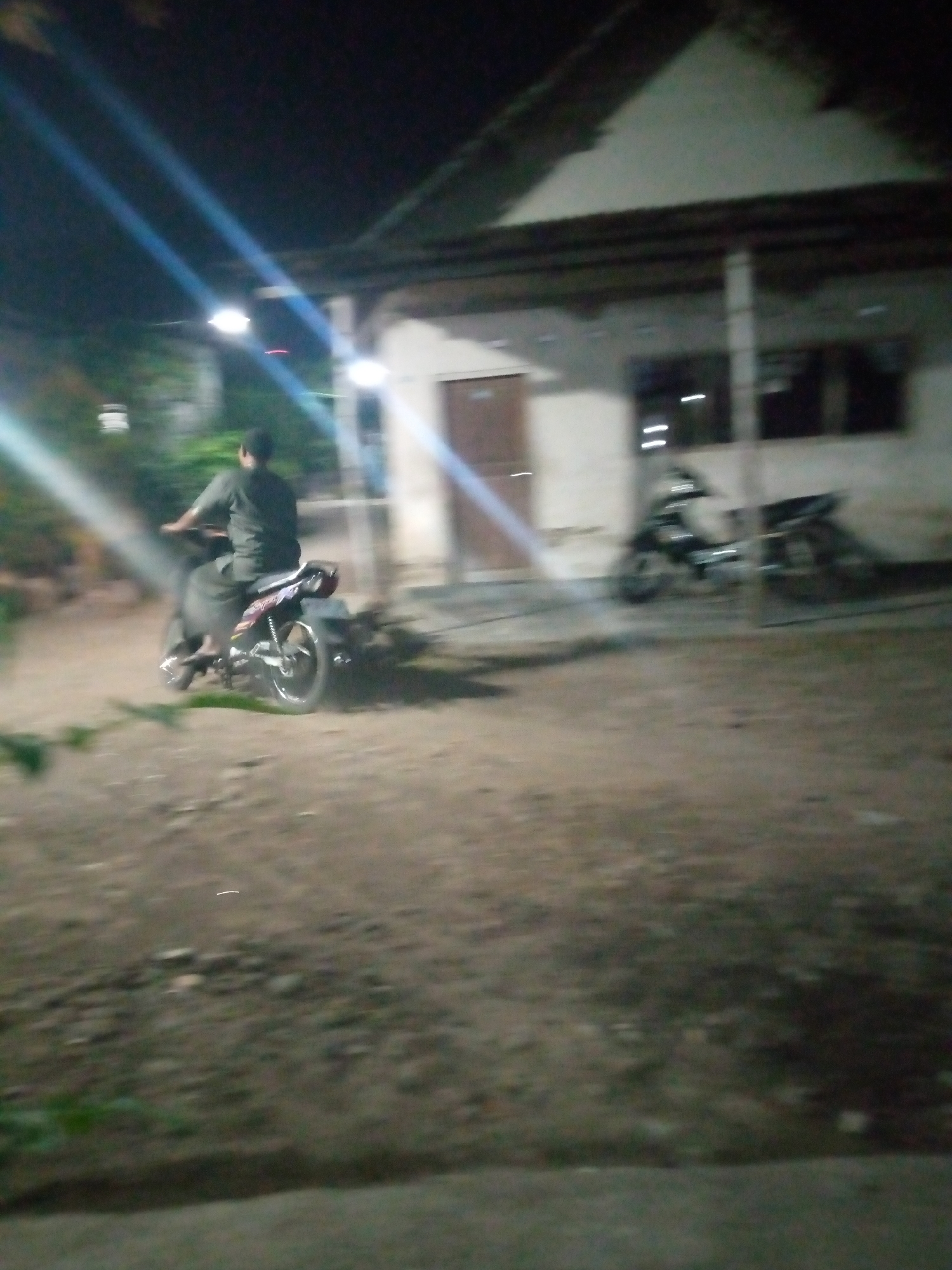 At night in Sumberwono, Indonesia, a person is riding a motorcycle. The motorcycle is dark colored with a red and white decal on the rear fender. The rider is wearing a dark shirt and pants. They are on a dirt or gravel road in front of a low-slung building with a dark roof and white walls. The building has several windows. Another motorcycle is parked to the right of the building. There are lights on the building and possibly from a street light. The scene is dimly lit and appears to be outdoors.