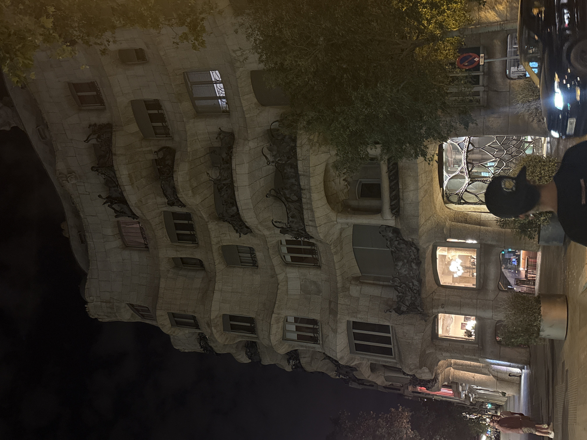 The image shows a night view of Casa Milà, also known as La Pedrera, in Barcelona, Spain. The building is a modernist architectural landmark designed by Antoni Gaudí. It features a distinctive undulating facade with stone balconies and wrought-iron decorations. The windows of the building are illuminated, revealing interior spaces. A person with a baseball cap is partially visible on the right, and a car is visible in the background. There is a street with people walking. The sky is dark, and trees are seen on the left.