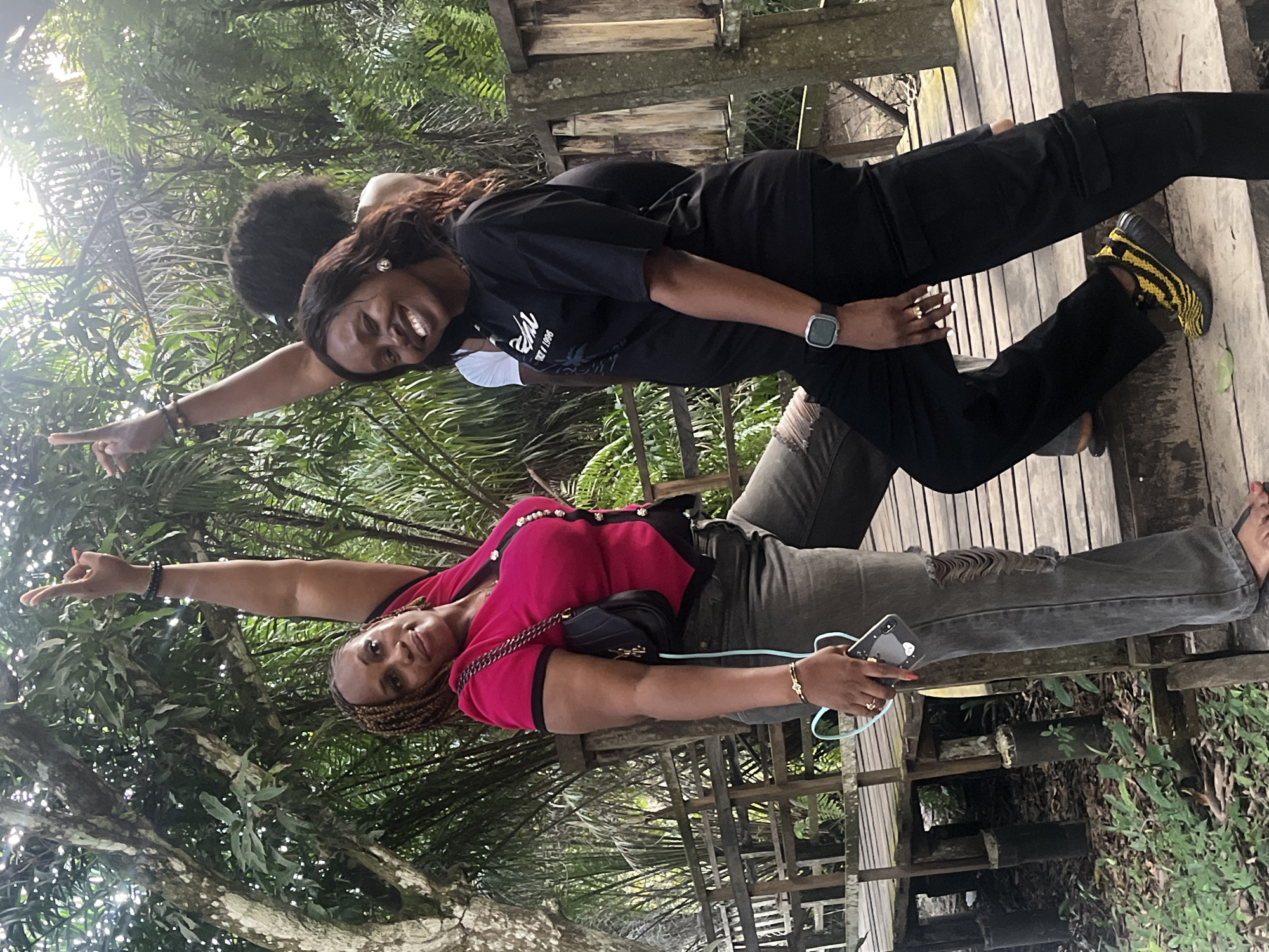 In Lekki, Nigeria, three individuals are captured in a candid outdoor moment, surrounded by lush greenery and wooden structures. Two women are prominently featured, one in the foreground wearing a vibrant pink and black top paired with dark, distressed jeans. She's holding a smartphone and has one arm raised, her fingers splayed in a playful gesture. Beside her, another woman in a black t-shirt and black cargo pants is smiling broadly, also with an arm raised. Behind them, a third person, whose face is not visible, has an arm raised in a similar pose. The setting appears to be a boardwalk or elevated walkway constructed from weathered wood, suggesting a natural or recreational area within Lekki. The foliage, a mix of trees and palms, indicates a tropical environment. The lighting suggests it's daytime, with sunlight filtering through the leaves.