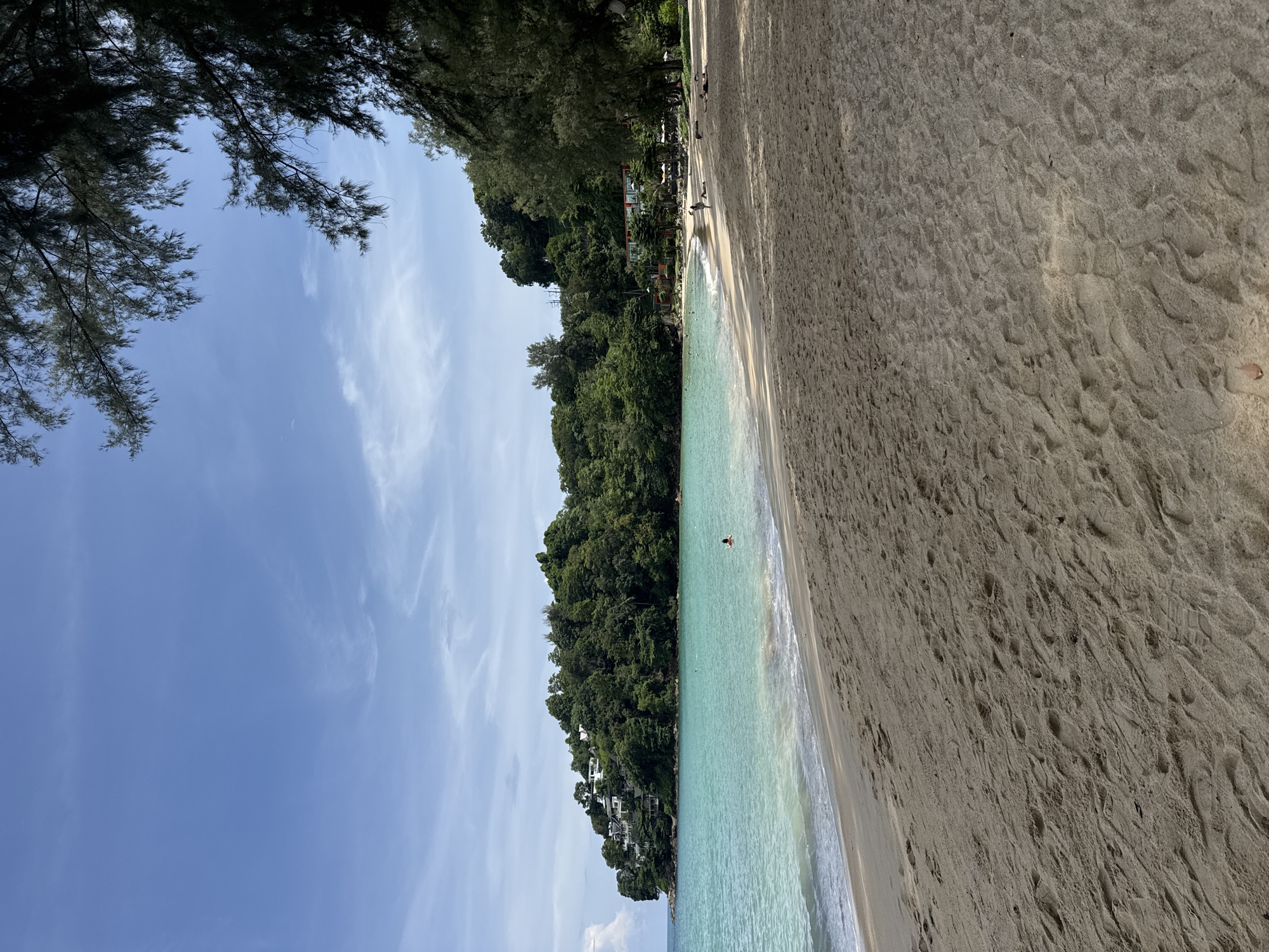 The image depicts a tropical beach scene in Ban Bang Thao, Thailand. The foreground is dominated by textured sand, showing numerous footprints. A gentle wave laps at the shore, separating the sand from the clear, turquoise ocean. In the mid-ground, the ocean extends towards a lush, green hillside covered in dense foliage and dotted with white buildings, suggesting a resort or residential area. A person can be seen swimming in the water. The background features a clear blue sky with scattered wispy clouds. To the right of the frame, the dark branches of pine trees overhang the scene, casting shadows on the sand. Several other people are visible on the beach and in the shallow water further down the coastline. The overall impression is one of a serene and picturesque beach environment.