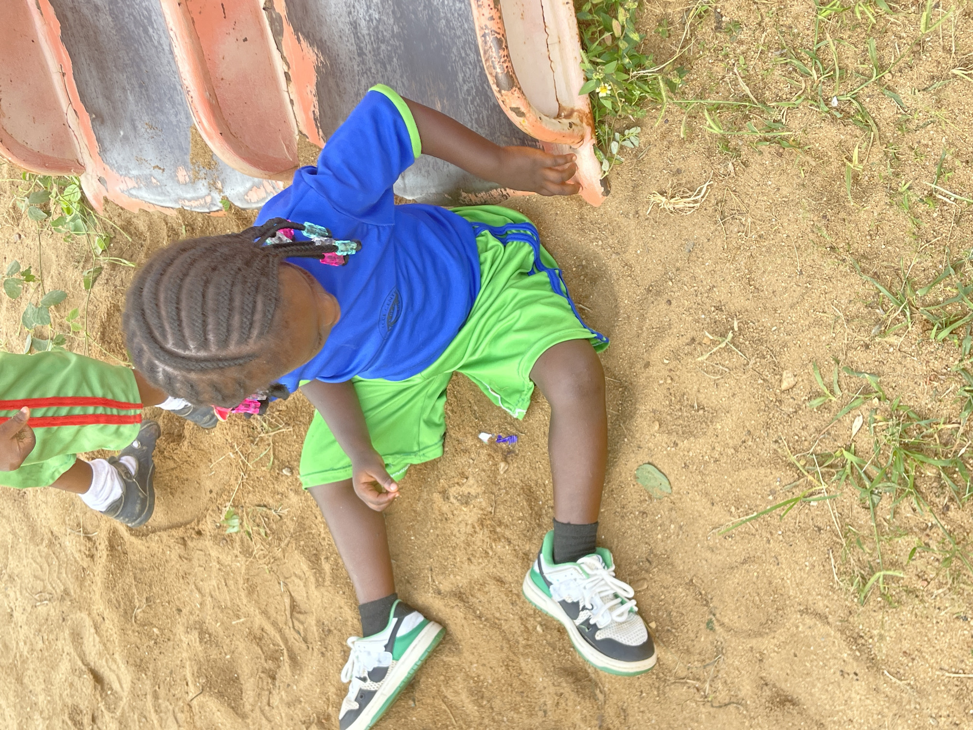 A child sits in the sand at the edge of a playground slide in Akwanga, Nigeria. The child wears a blue shirt with green trim, green shorts with blue trim, and black socks with green and white shoes. The child has dark hair with multicolored barrettes and rests one hand on the slide. Another child, wearing a green outfit with red trim, is partially visible on the left. The sand is mixed with grass and debris.