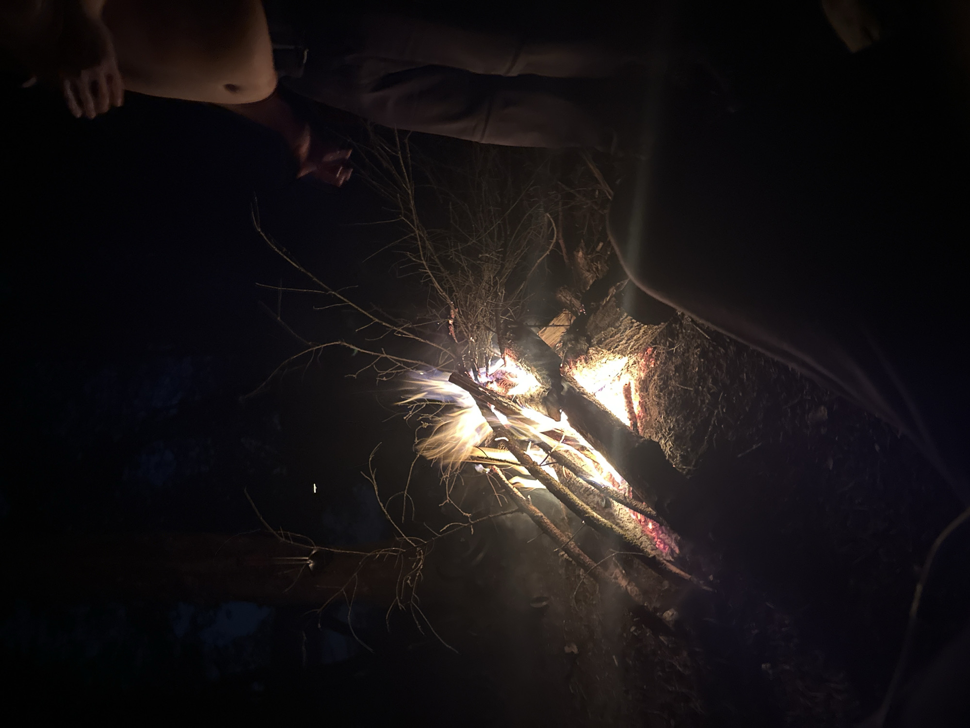 A night scene in what appears to be a rural location, possibly in Папярнянскі сельскі Савет, Belarus, features a small campfire. The flames are primarily white and yellow, consuming several thin branches and small logs. The fire is situated on the ground. Three people are partially visible in the frame, appearing to be sitting around the fire. The environment is dark, with limited light beyond that cast by the flames. The ground appears to be covered with dirt and possibly some vegetation. Some bare branches are also visible near the fire.