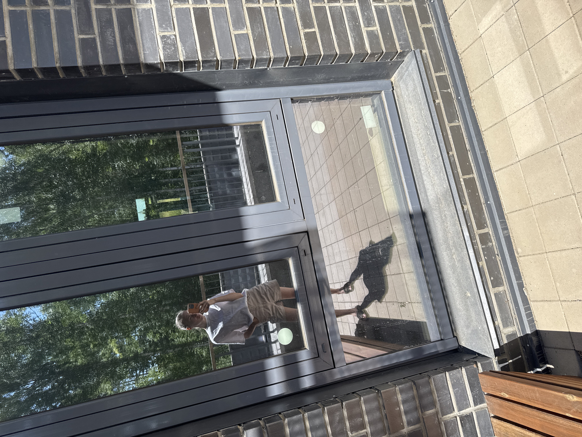 The image shows a reflection of a young woman taking a selfie in a large window. She is wearing a white t-shirt and light-colored shorts, with her hair pulled back. The reflection shows her holding a smartphone. The window is part of a modern building, with dark gray framing and dark brick exterior walls. In the reflection, trees and a railing are visible in the background, suggesting an outdoor setting, possibly a balcony or patio. The ground in front of the window is paved with light-colored tiles. A wooden bench is partially visible in the lower left corner of the frame, outside the window. The scene appears to be in Krasnoyarsk, Russia, based on the provided context.