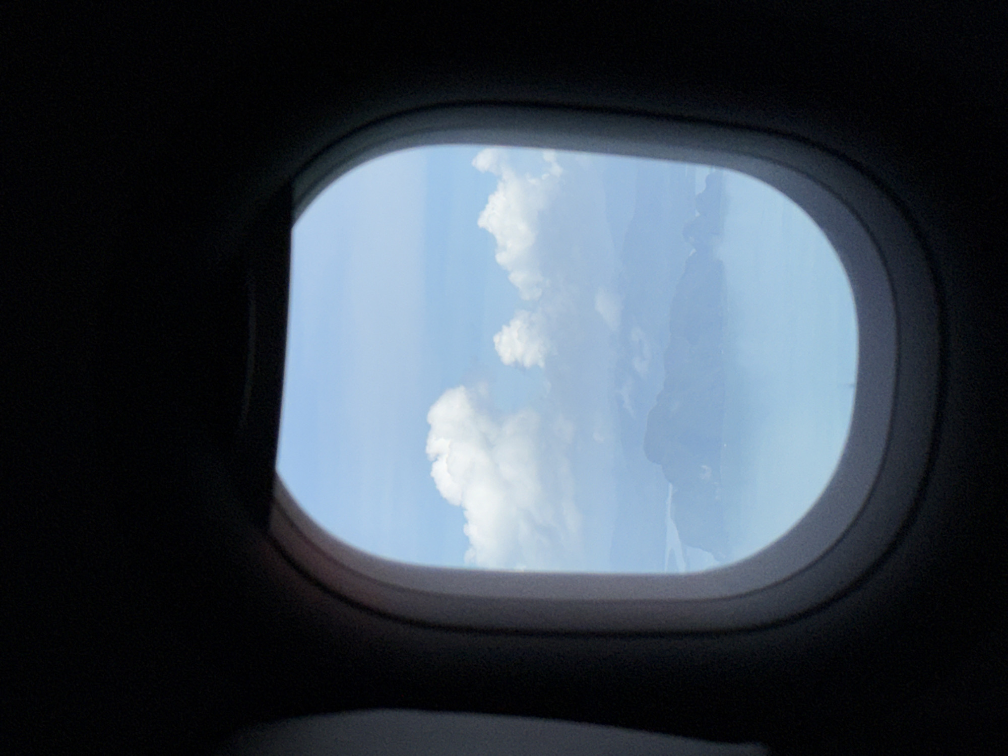 The image is taken from an airplane window, looking out at a hazy sky and islands in the distance. The window frame, which is dark, creates a circular view of the outside. The sky is a light blue, with scattered white clouds. Below the clouds, islands are visible, surrounded by a calm, light blue body of water, likely the Andaman Sea. The islands are mountainous and appear to be covered in lush greenery, though the haze softens their details. The overall impression is one of a serene aerial landscape. While the exact location within Ban Khao Thong, Thailand cannot be definitively identified from this image alone due to the distance and atmospheric conditions, the scenery is consistent with the geography of coastal Thailand, particularly areas known for their karst formations and islands like those found in Phang Nga Bay, which is near Ban Khao Thong. There are no people or specific objects visible in the scene, besides the natural elements of sky, clouds, water, and landforms. The focus is on the expansive and tranquil view from the airplane.