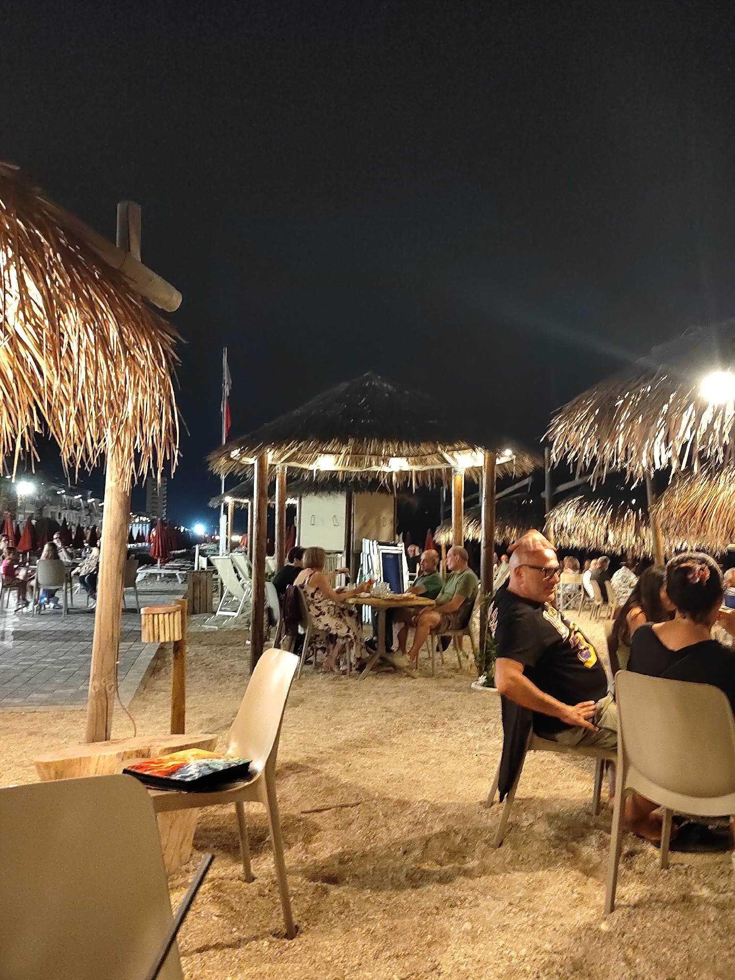 This is a night-time photograph of an outdoor beachside dining area in Porto Recanati, Italy. The scene is illuminated by artificial lighting and depicts several thatched-roof umbrellas providing shade over tables and chairs. In the foreground, a man with glasses and a black t-shirt is seated at a table, looking to his right. To his right, a woman with dark hair is also seated. In the middle ground, a group of people are seated at a table under one of the umbrellas, appearing to be dining. Further back, other patrons and beach chairs are visible, along with red umbrellas. The ground is covered in sand and small stones. The overall ambiance is that of a lively evening at a beach restaurant.