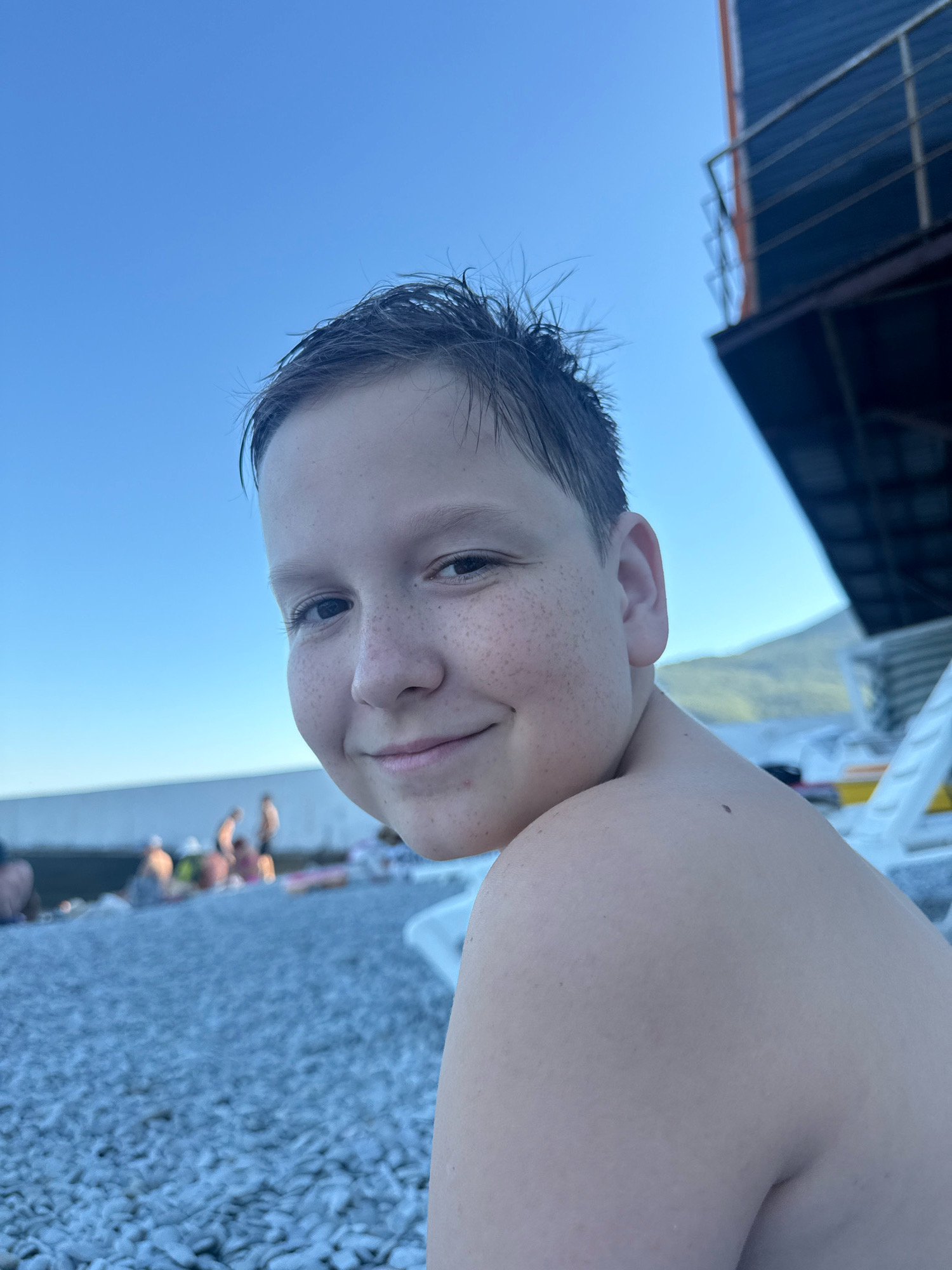 A medium-shot, eye-level, candid portrait captures a young boy smiling as he sits on a pebbled beach under a clear blue sky. The boy, with dark, wet hair and freckles across his nose and cheeks, faces the camera and is cropped from the shoulders up, with his left shoulder prominently in the foreground. In the background, out of focus, are beachgoers, lounge chairs, and a dark, tiered structure with railings, possibly a building or boat, extending from the top right corner. The beach stretches into the distance, transitioning to a body of water and then hills under the bright sky. The scene is consistent with a daytime setting on a beach in Partenit, Ukraine.