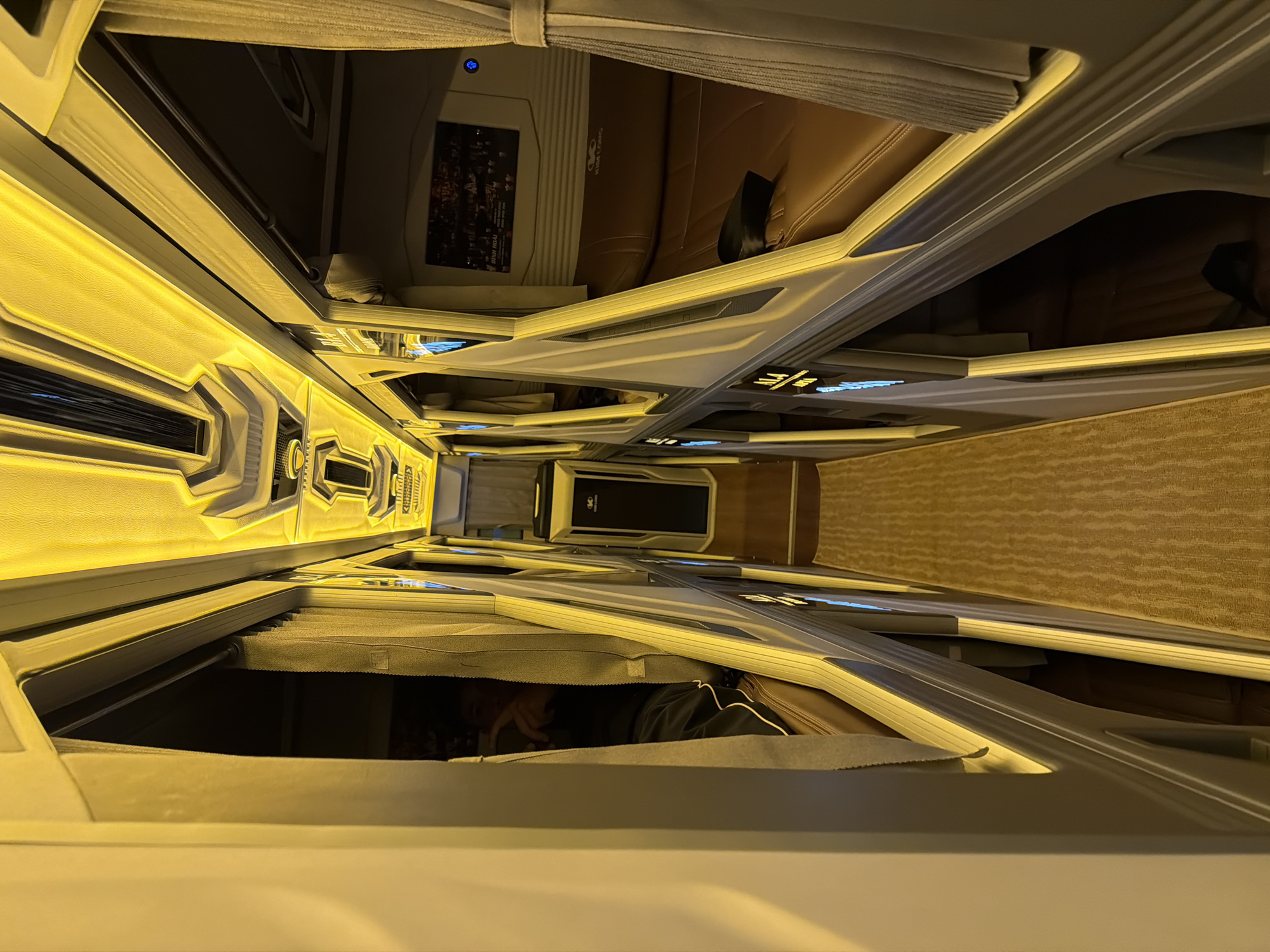 The image displays the interior of a modern sleeper bus, likely operating in Đà Nẵng, Vietnam, characterized by a series of capsule-like sleeping pods. The perspective is a top-down view looking along a narrow aisle lined with these individual berths, with the image rotated clockwise.
Warm, golden LED strip lighting illuminates the interior of each pod and along the aisle, creating a cozy atmosphere. The pods feature contemporary design elements with grey and white structured panels. Many pods are equipped with brown upholstered seats or beds. In one of the upper pods, a screen is visible, displaying content with Vietnamese text that includes "ĐÊM THU HÀ NỘI" (Hanoi Autumn Night). The logo "King Long," a common bus manufacturer, is also discernable near the screen. This pod also shows a black seatbelt.
Further down the aisle, in a lower pod, a person is partially visible, appearing to be resting or engaging with a device within their private space. They are wearing dark clothing with white stripes. The floor of the aisle is covered with a light brown, patterned carpet. Small blue indicator lights are intermittently visible on the pod structures. The scene suggests an enclosed environment, typical for overnight or long-distance travel, likely during the night due to the prominent artificial lighting.