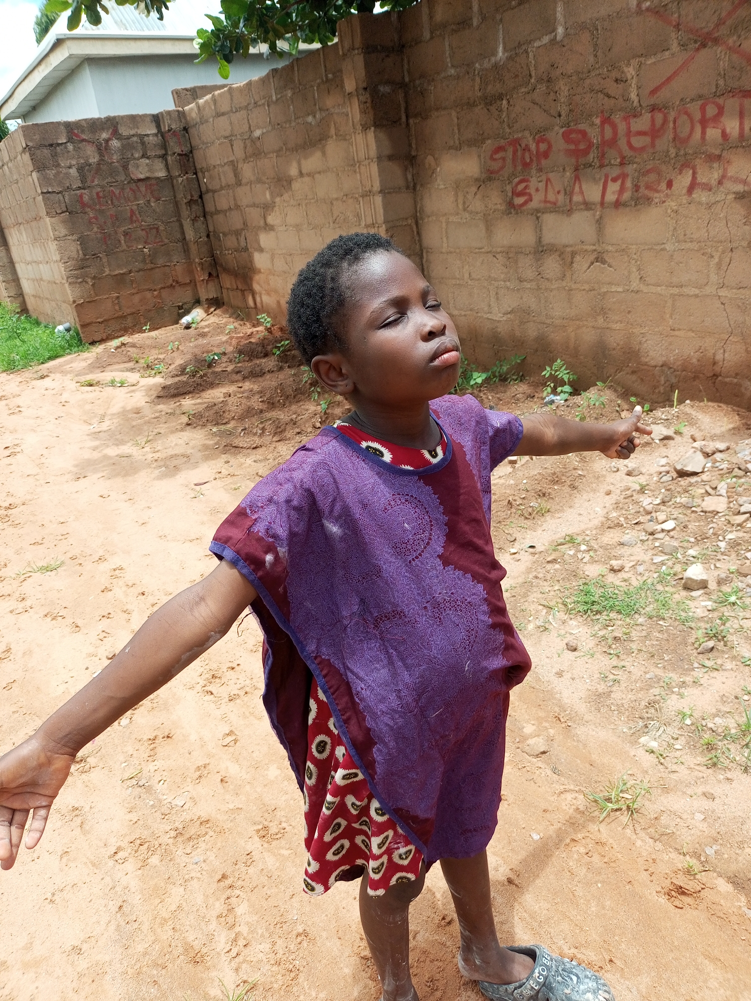 In a sandy, outdoor environment, likely within the city of Tempure, Nigeria, a young child stands with their arms outstretched and eyes closed, appearing to be praying or basking in the sun. The child is wearing a purple, patterned tunic over a red and white patterned skirt. Behind the child, a rough brick wall is visible, marked with red graffiti. The ground is sandy and uneven, with sparse vegetation and scattered rocks. The lighting suggests it is daytime.
