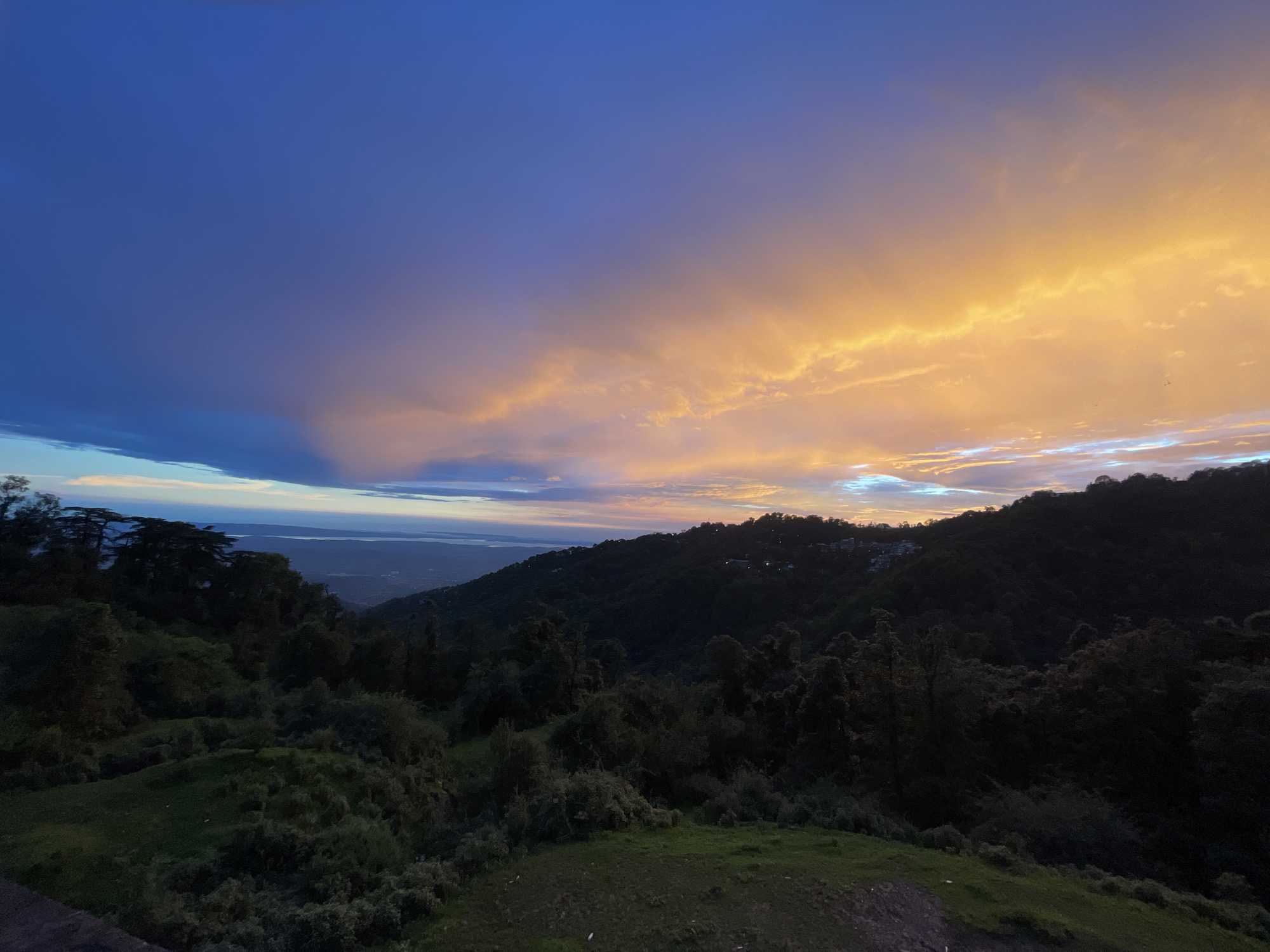 This image depicts a serene sunset over a hilly landscape in Dharamkot, India. The sky is a dramatic canvas of colors, transitioning from deep blue at the top to vibrant oranges and yellows near the horizon, with wisps of clouds catching the last rays of sunlight. Below, the verdant hills are covered in dense vegetation and scattered trees, casting long shadows in the fading light. A small village or settlement is visible nestled on the slopes of the furthest hill, its lights beginning to twinkle. The foreground features a grassy slope with some rocks and dirt, suggesting a viewpoint overlooking the valley. The overall atmosphere is peaceful and tranquil, capturing the beauty of nature at dusk.