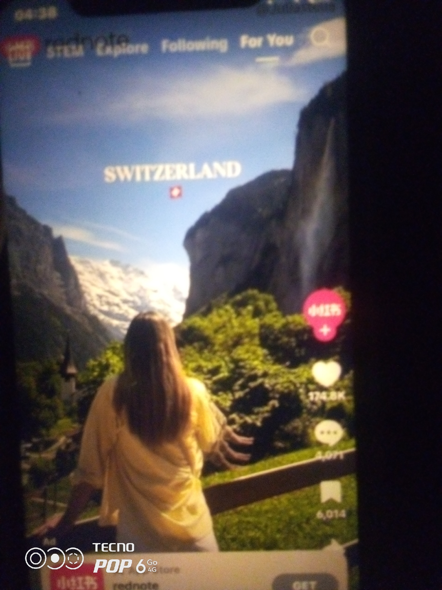 The image shows a person from behind, wearing a yellow shirt and white pants, standing on a balcony or railing overlooking a scenic mountain landscape with a waterfall. The person is facing away from the camera, with their long, brown hair visible. The background features snow-capped mountains and lush green trees. Text overlays on the screen indicate the location as "SWITZERLAND" and also display various interface elements likely from a social media or video app, including likes, comments, and share counts. There are also logos and branding, including "TECNO POP 6 Go 4G". The context of Howayi, Nigeria is not visually present in the image itself.