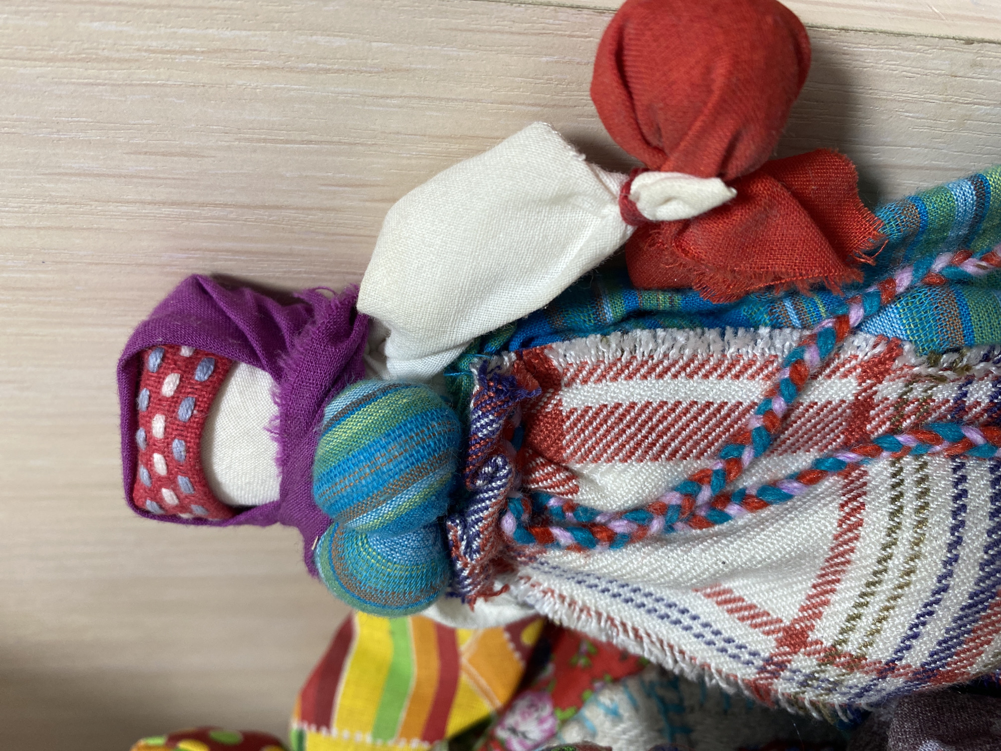 A close-up shot shows a handcrafted fabric doll against a light wooden background. The doll's head is covered with a red fabric, suggesting a scarf or headdress. The doll features colorful patchwork fabric clothing, including a purple headscarf with white and red polka dots and a plaid dress with red, white, and blue patterns. The head and body are made of various materials, including white fabric, creating a layered effect. The doll's colorful appearance and handcrafted design indicate its origin as a traditional folk toy or craft. The photograph's composition emphasizes the details and textures of the doll's construction, with no people or specific location visible in the frame. The doll's position and orientation are not indicative of the city of Noviy, Russia.
