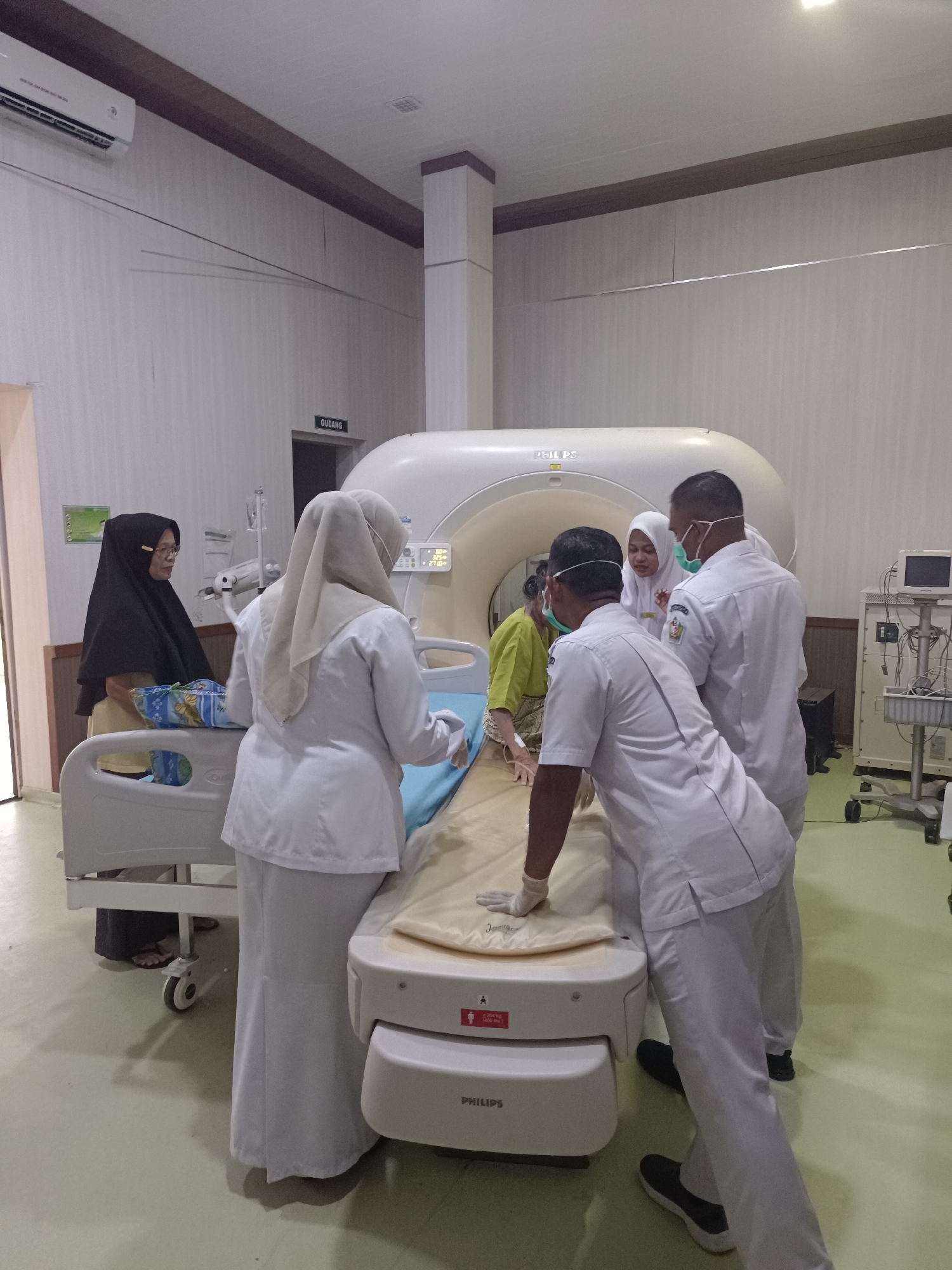 In a well-lit medical facility, likely a hospital radiology department, a patient is being prepared for a CT scan. The patient, wearing a lime green top, is lying on a medical bed adjacent to a large, white CT scanner machine. A healthcare professional, wearing a white hijab and scrub suit, stands on the left side of the bed, attending to the patient. Another healthcare professional, in a white scrub suit and face mask, is positioned on the right side of the bed, adjusting something on the patient's arm. A third healthcare professional, also in a white scrub suit and face mask, is visible behind them, appearing to supervise or assist. In the background, a sign indicates "GUDANG," which translates to "STORAGE" in English, suggesting a utilitarian area within the facility. The environment is clean and functional, typical of a medical setting, with an air conditioning unit mounted high on the wall. The overall scene depicts a clinical procedure in progress, with medical staff attending to a patient. The location is identified as Mesjid Reuntoh, Indonesia.