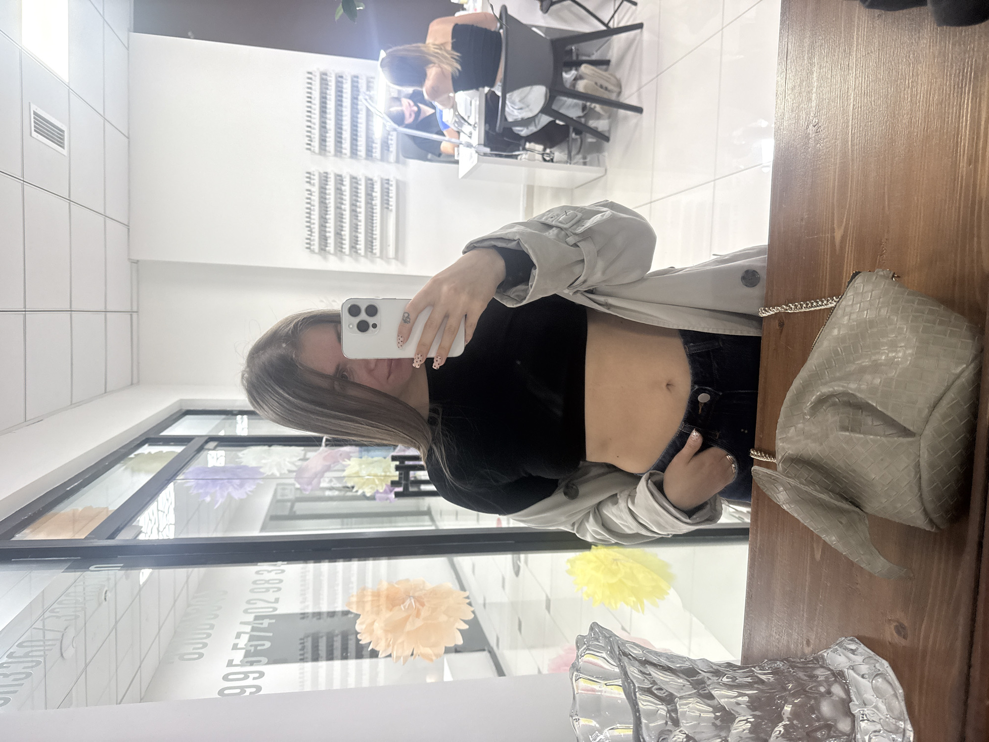 A woman is taking a mirror selfie in a nail salon in Batumi, Georgia. She is wearing a black crop top, jeans, and a light grey trench coat. The woman is holding a smartphone with a white case, showcasing decorated nails. A beige woven handbag with a chain strap rests on a wooden surface. In the background, another woman is receiving a manicure. The salon features white walls, a nail polish display, and decorative paper flowers in the window. The phone number 995-574-02-98-31 is visible on the window. The environment suggests a brightly lit and modern salon setting.