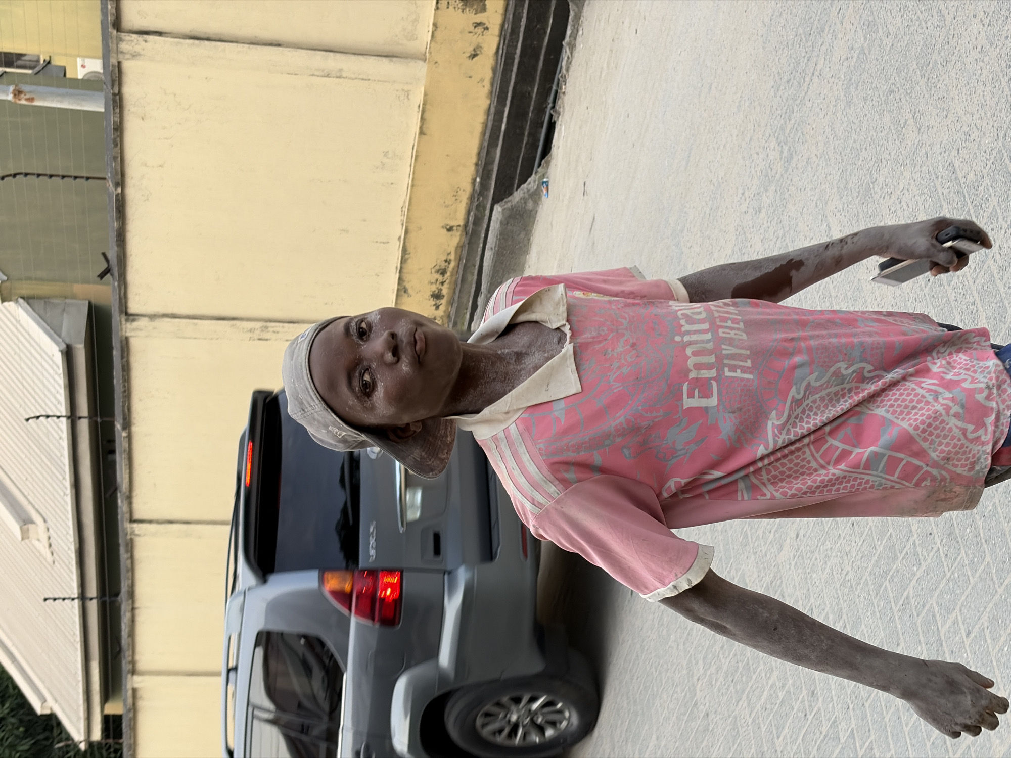 This image, taken in Igboefon, Nigeria, features a young person, likely male, standing outdoors. The individual has dark skin and is covered in a significant amount of light-colored dust or powder, particularly visible on their face, arms, and clothes. They are wearing a pink short-sleeved t-shirt with grey patterns, featuring the "Emirates" logo and partial text "FLY BETT" (likely "FLY BETTER"), along with Adidas's three-stripe design on the shoulders. A grey cap is worn on their head. In their right hand, they hold a dark-colored smartphone.
The person stands with a neutral expression, looking directly at the camera. The background shows a light grey SUV parked behind them, with its rear brake lights visible. Beyond the vehicle, there are buildings with pale yellow walls and a white-roofed structure to the left. The ground is paved, possibly concrete, and also appears covered in dust, consistent with the person's appearance.
The scene is brightly lit, suggesting clear daylight conditions, possibly midday or afternoon. The extensive dust suggests an environment where activities like construction, demolition, or dry earthwork are taking place, or the person has been involved in such work. The overall setting appears to be an urban or semi-urban area.