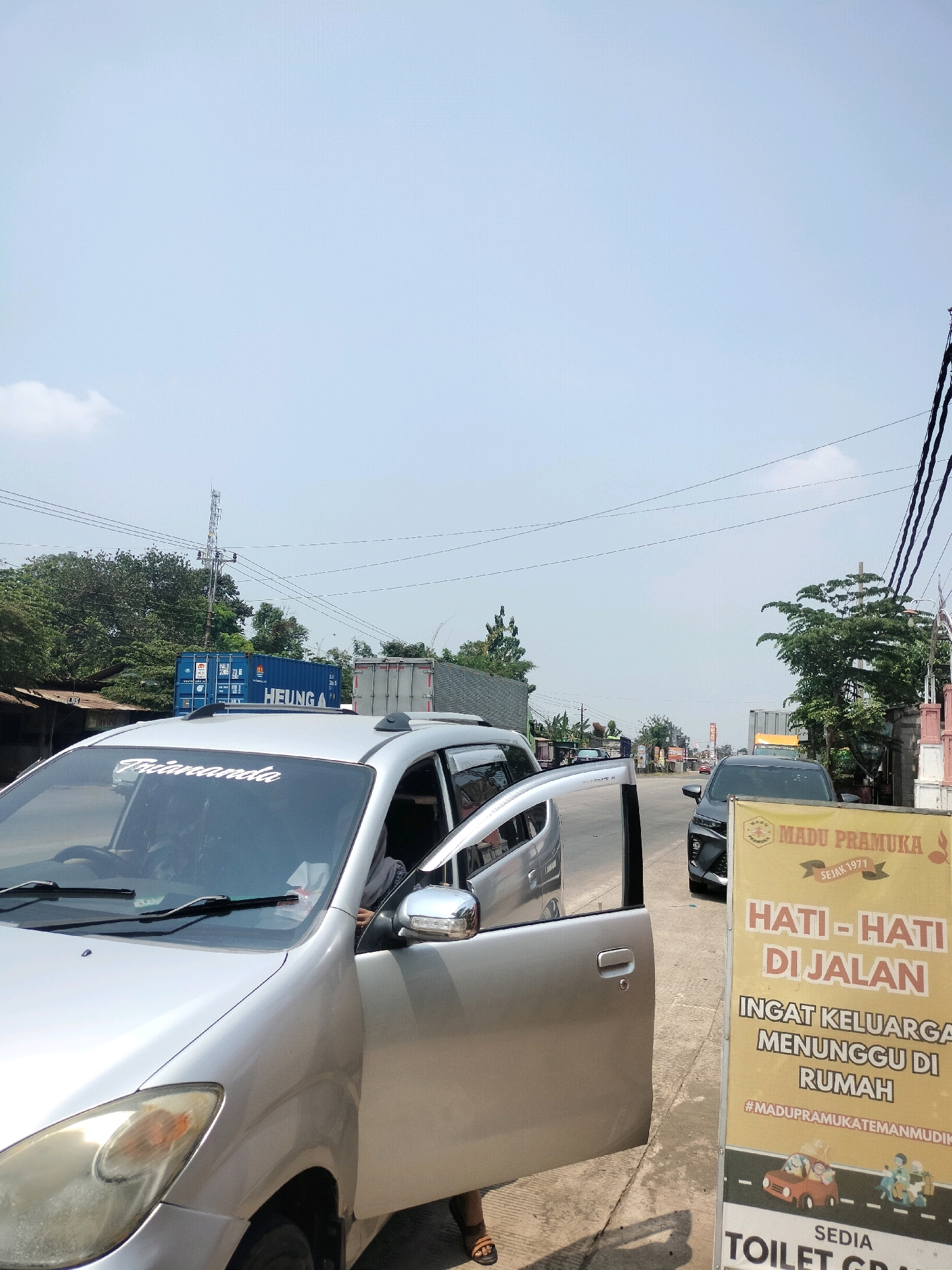 The image shows a roadside scene in Kutosari, Indonesia, on a sunny day. A silver MPV with its driver-side door open is parked on the right side of a paved road. A person, wearing sandals, is partially visible exiting the vehicle. To the right of the car, a large yellow banner with text in Indonesian is displayed. The banner reads "MADU PRAMUKA SEJAK 1971 HATI-HATI DI JALAN INGAT KELUARGA MENUNGGU DI RUMAH #MADUPRAMUKATEMANMUDIK SEDIA TOILET GRATIS". This translates to "Pramuka Honey Since 1971 Be Careful on the Road Remember Family Waiting at Home #PramukaFriendsOnMudik Free Toilet Available". The background features trees, electrical poles with wires, and several large shipping containers, including one blue container labeled "HEUNG A". A grey truck is also visible. The sky is clear blue with a few scattered clouds.