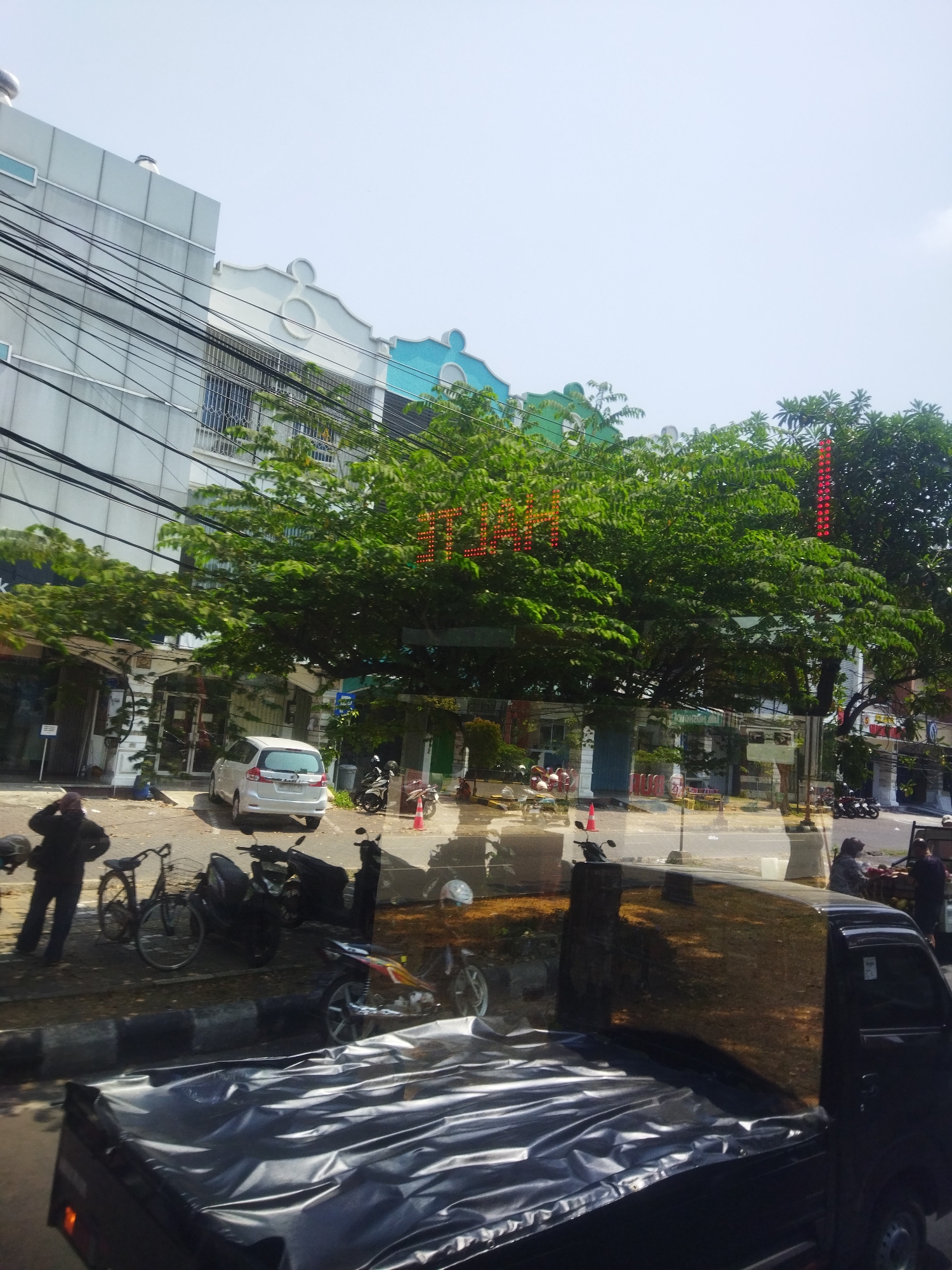 The image depicts a street scene in Grogol, Indonesia, with several buildings, vehicles, and people. A white minivan is parked on the street in front of a building, while several motorcycles and a bicycle are also visible. A person is standing next to the bicycle, and another person is seen in the reflection of a vehicle. The buildings have a modern architectural style, with clean lines and some decorative elements. One building has a large tree in front of it, with red LED lights forming a pattern on the branches. The street appears to be a commercial area, with shops or businesses lining the road. The sky is clear and bright, suggesting a sunny day. The image seems to have been taken from inside a vehicle, possibly a car, due to the perspective and the reflection on the glass.