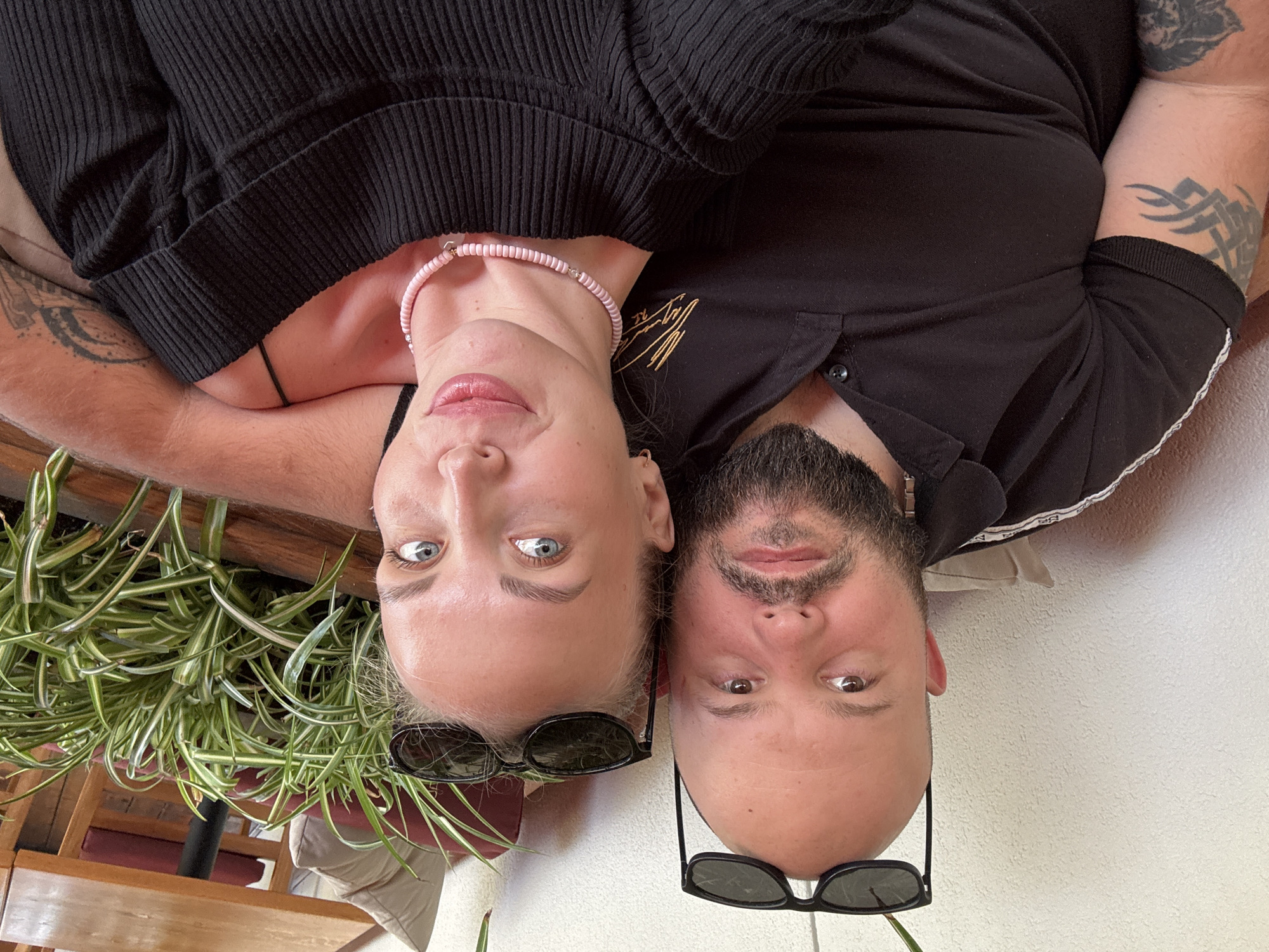 An overhead photograph captures a man and a woman lying side-by-side, their heads close together, looking directly up at the camera. The image is rotated 90 degrees clockwise.
The woman, on the left, has blonde hair, blue eyes, and light skin. She is wearing a black ribbed top and a pink beaded choker necklace with a small metallic accent. Her lips are slightly parted, revealing a touch of pink lipstick. Sunglasses are pushed onto her forehead. A tattoo is visible on her left arm.
The man, on the right, has dark hair, brown eyes, a full beard, and light skin. He is wearing a black polo shirt with a gold embroidered logo on the left chest and a white striped trim on the sleeve, which has indistinct repeating text. Sunglasses are also pushed onto his forehead. Tattoos, including a tribal-style design, are visible on his right arm.
Both subjects have neutral, relaxed expressions. Behind the woman, green, leafy plants and wooden furniture are visible. Behind the man is a light-colored, textured wall. The scene suggests a casual setting, possibly a cafe or an outdoor patio area in Sochi, Russia. The gold embroidered text on the man's shirt is illegible.