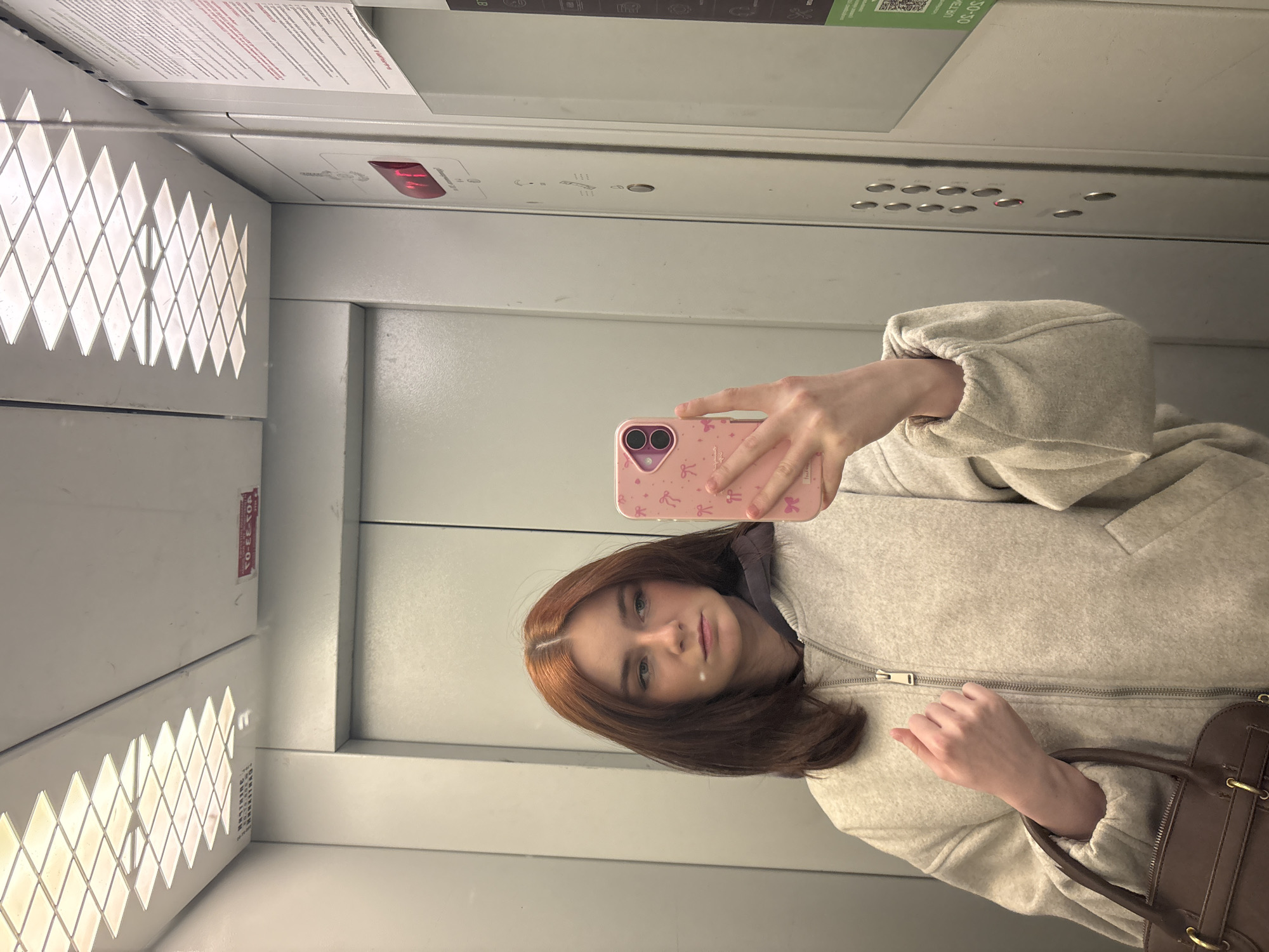 A person is taking a selfie in an elevator. The elevator's interior is gray and white, with a patterned ventilation grille visible. The person is holding a pink phone with decorative prints. The person has shoulder-length auburn hair and is wearing a light-colored jacket. A brown handbag is partially visible in the frame. A sign is at the top of the elevator. There are floor selection buttons with a light indicating which floor is selected. The location is the city of Unknown, Russia.